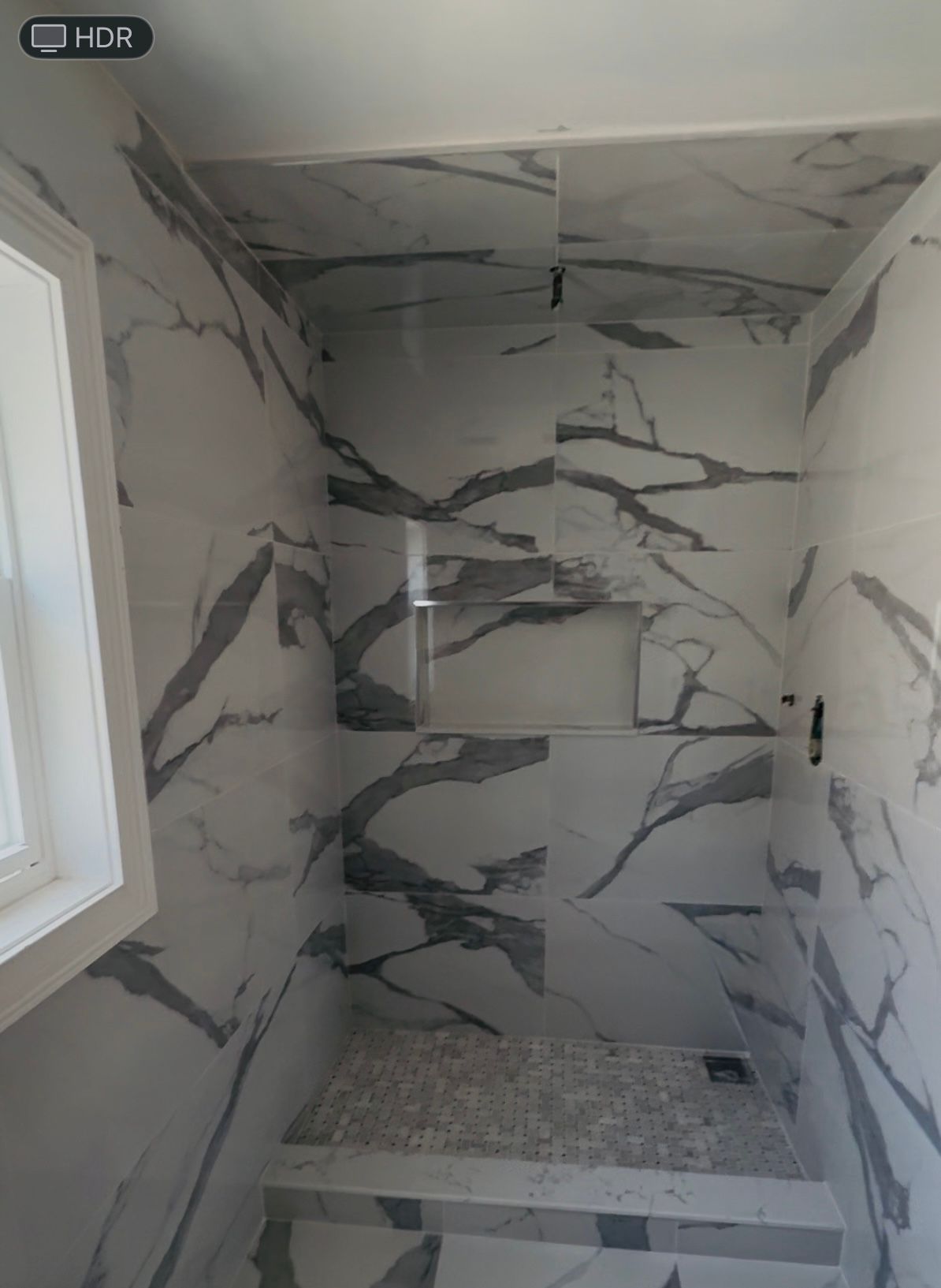 Shower with marble-look tiles, small recessed shelf, and pebble floor. A window is on the left.