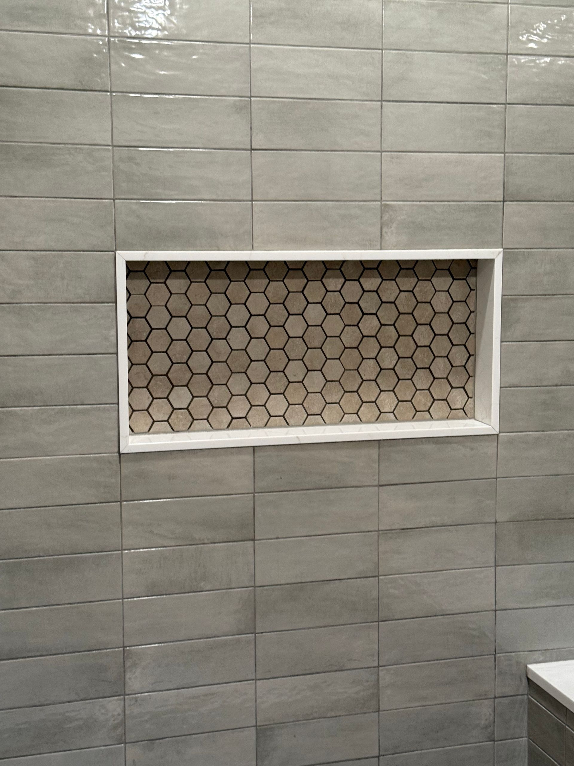 Gray tiled shower wall with recessed shelf filled with honeycomb-patterned tile, bordered by a white frame.