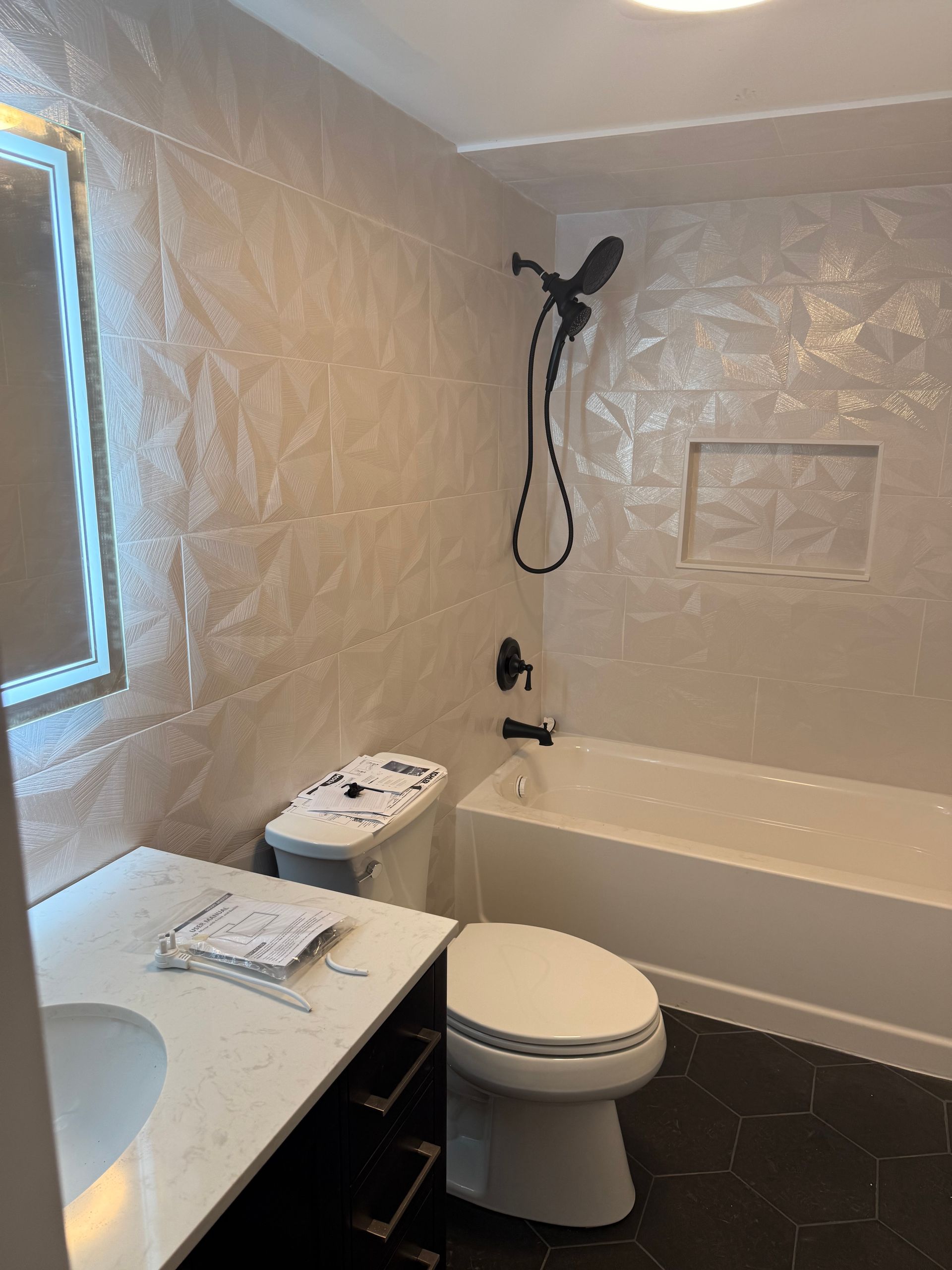 Bathroom with white geometric tiles, black fixtures, white tub, toilet, and vanity.