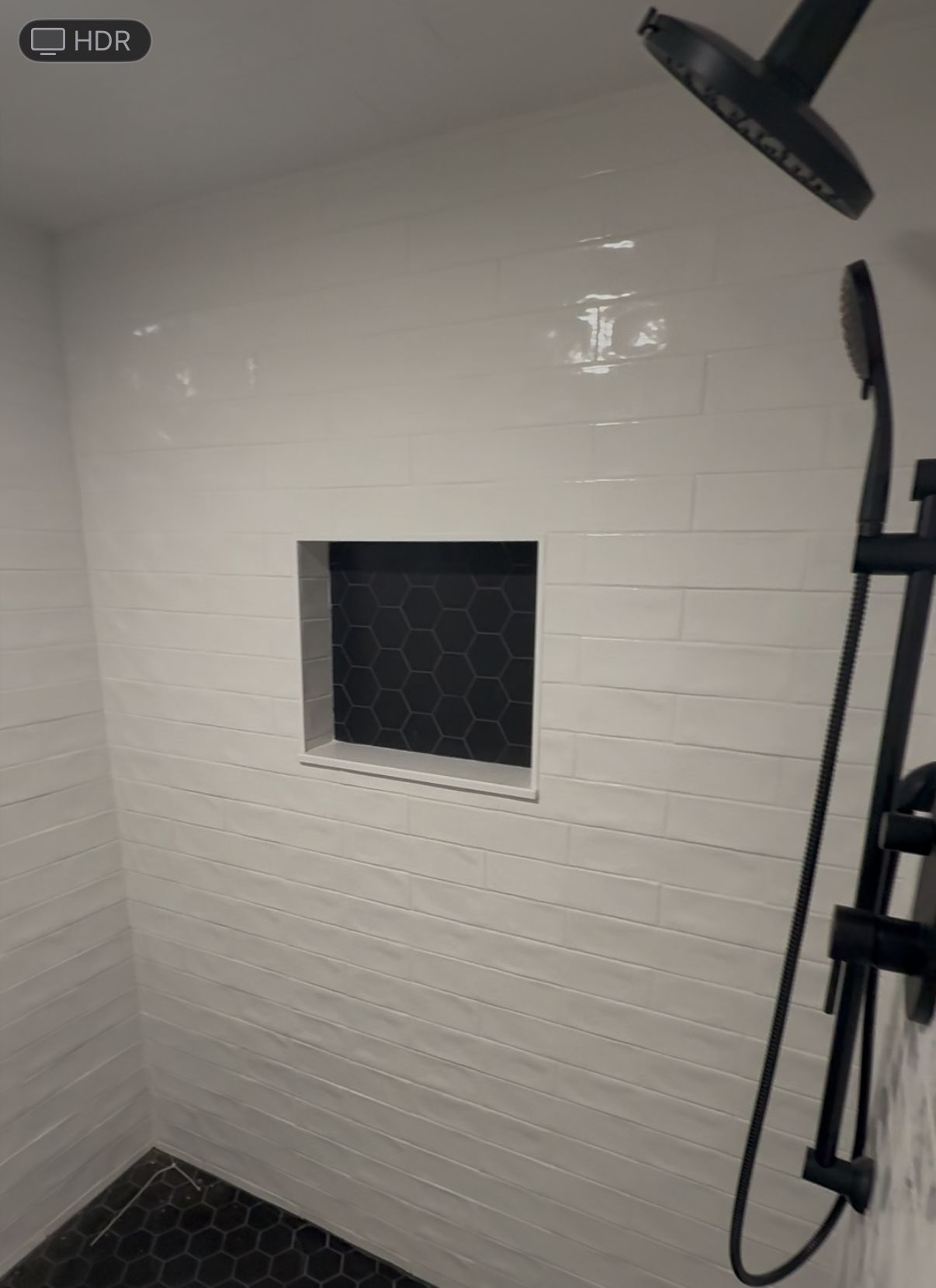 White-tiled shower with a black hexagon-tiled recessed shelf. Black showerhead and handheld shower.
