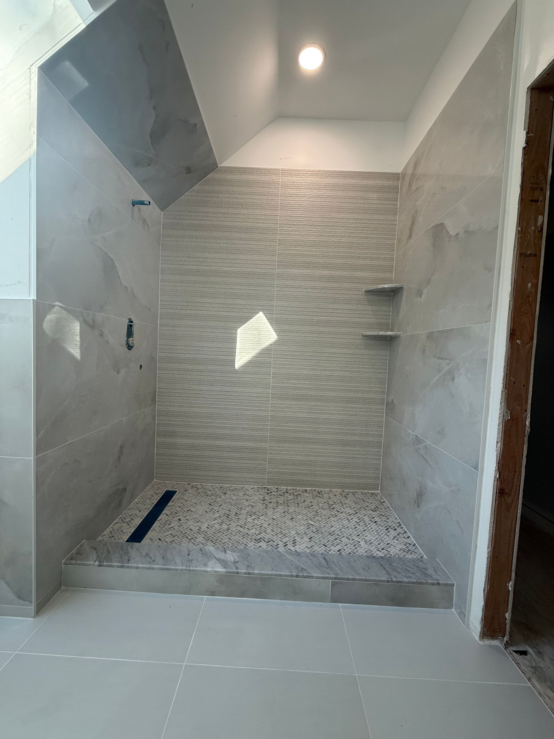 Newly constructed walk-in shower with gray tiles and stone floor; two wall shelves and sloped ceiling.