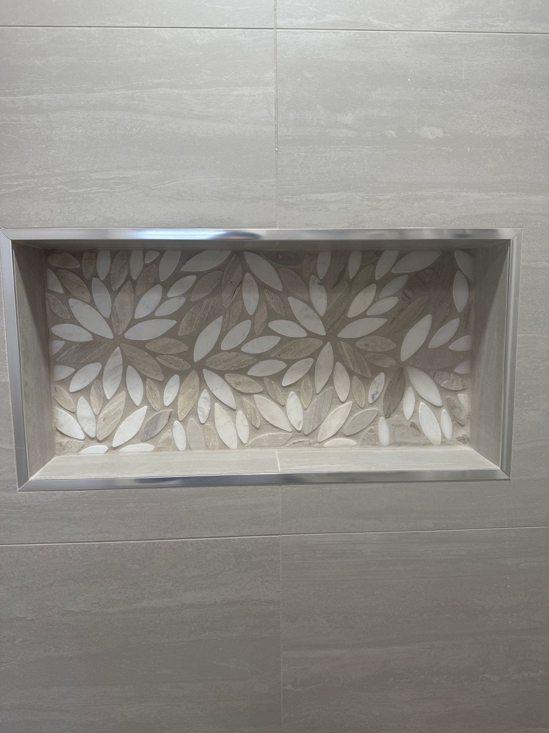 Recessed shower niche with leaf-patterned mosaic tile and metal trim, surrounded by light gray tiles.