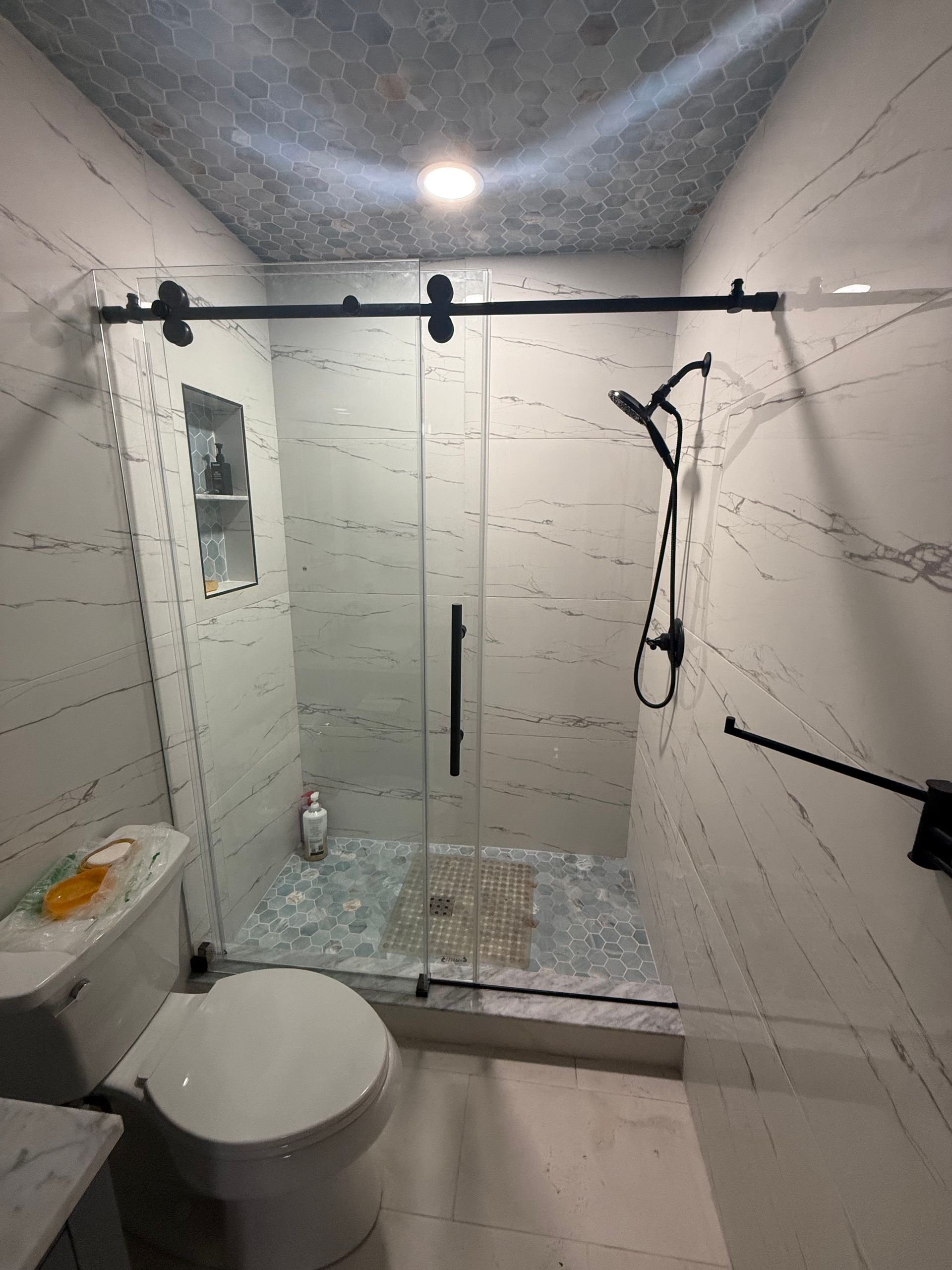 Modern bathroom with sliding glass shower door, marble-look walls, and black fixtures.