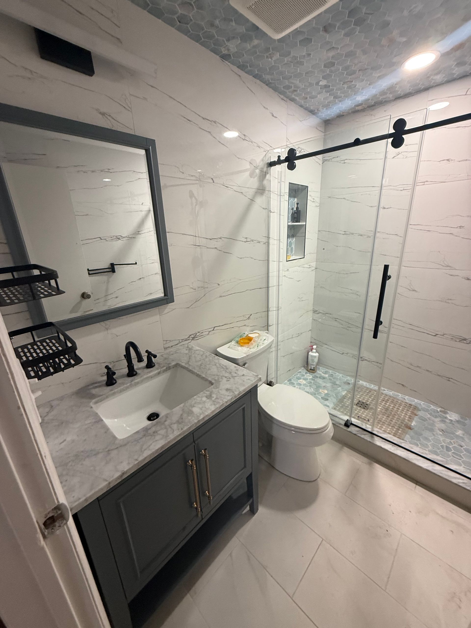 Modern bathroom with gray vanity, white marble walls, glass shower, and patterned ceiling.
