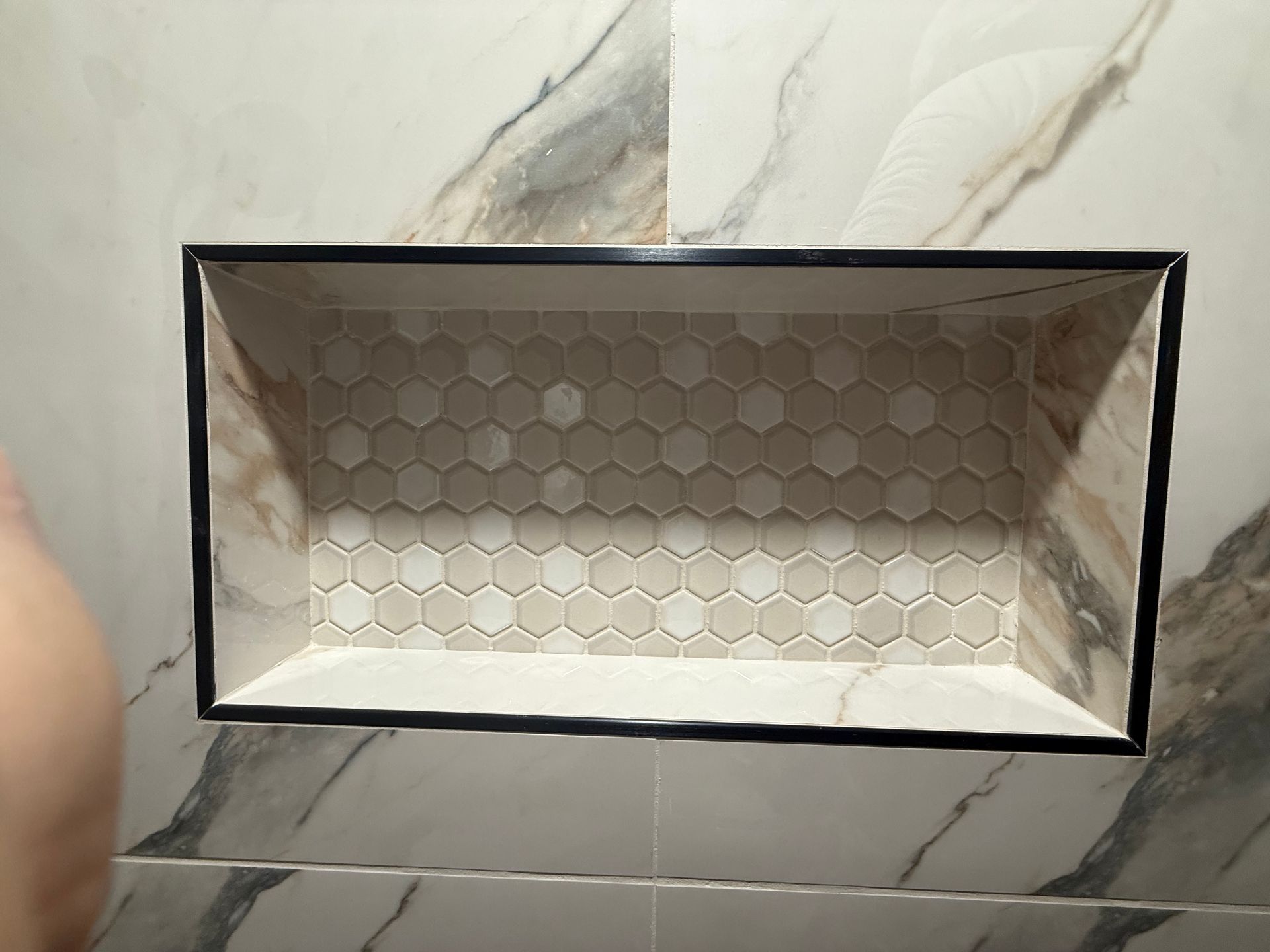 Built-in shower niche with hexagon tile backing, framed in black, set within marble-patterned tile walls.