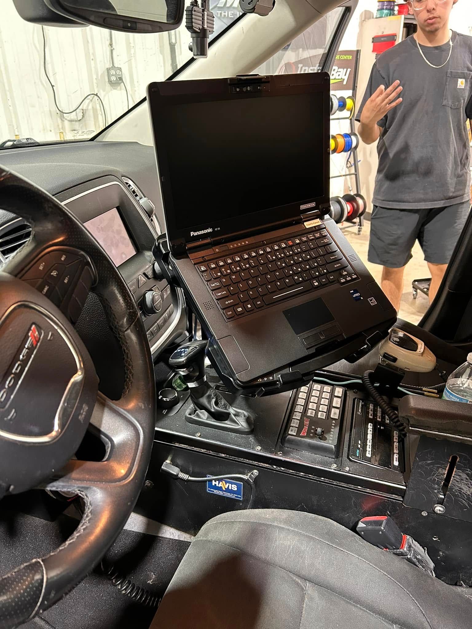 Interior of a police vehicle with laptop mounted on center console; person standing nearby.