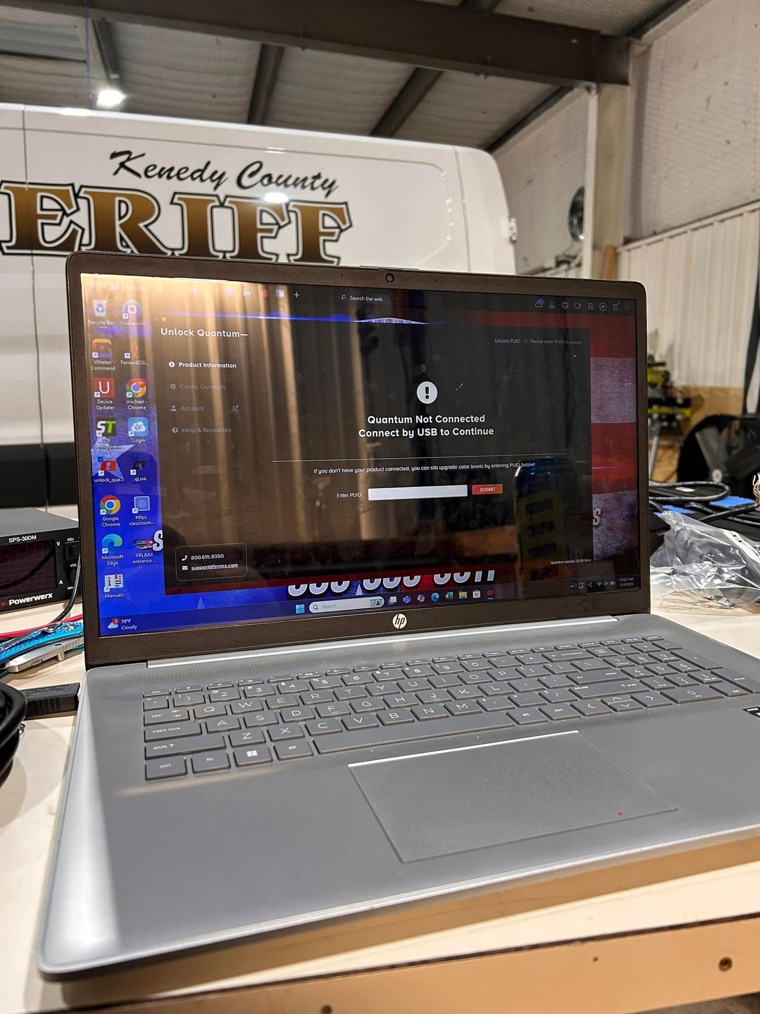 Silver laptop with cracked screen open on a wooden surface, partially obscuring a Kennedy County Sheriff vehicle.