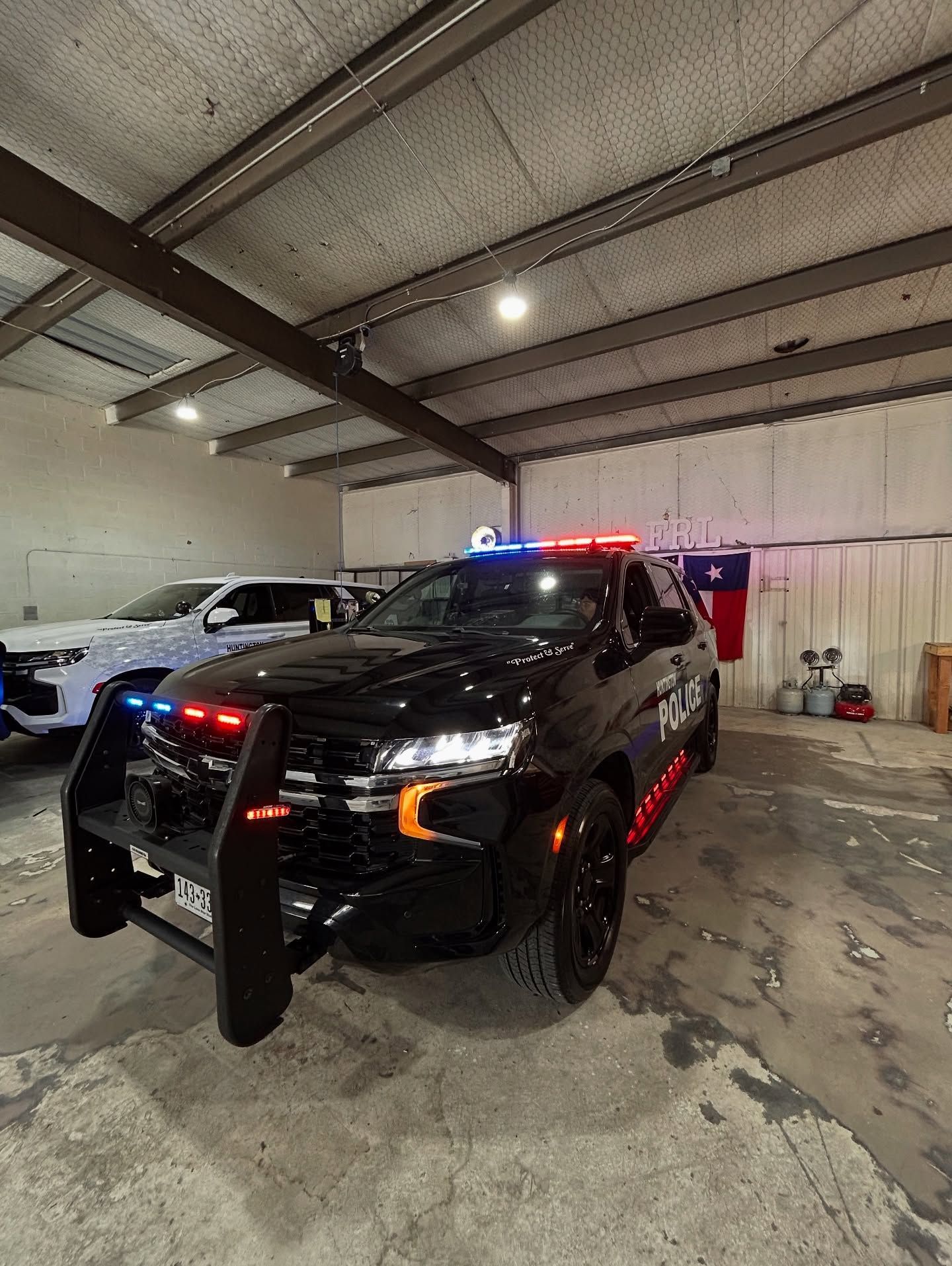 Black police SUV with emergency lights, parked in a garage. Another vehicle is partially visible on the left.