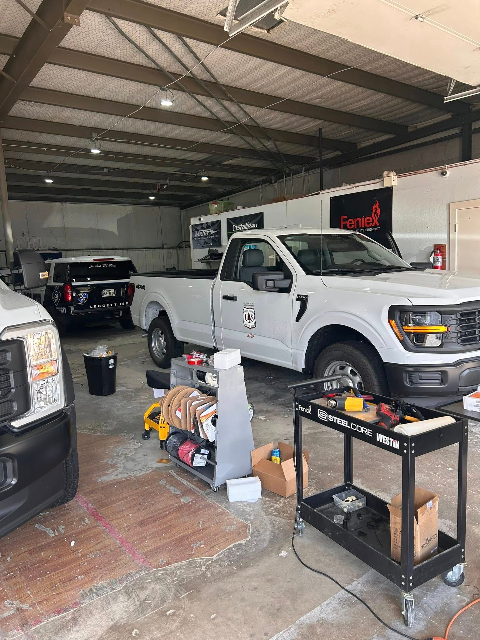 Inside a workshop, white work trucks are being worked on. Tools and equipment are scattered about.