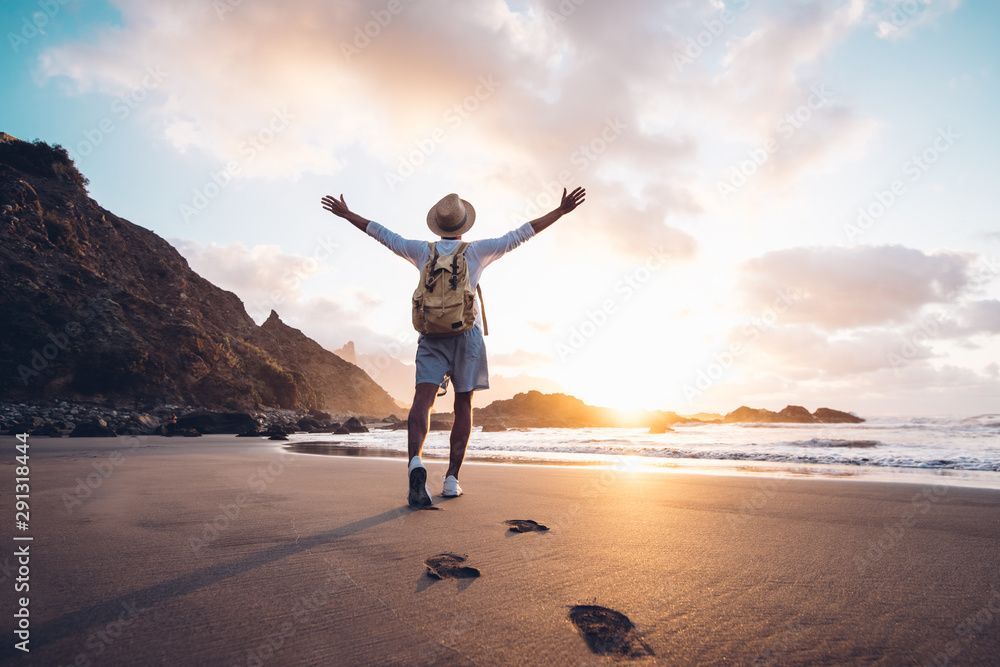 Person with arms outstretched on a beach at sunset, facing the ocean; backpack and hat.
