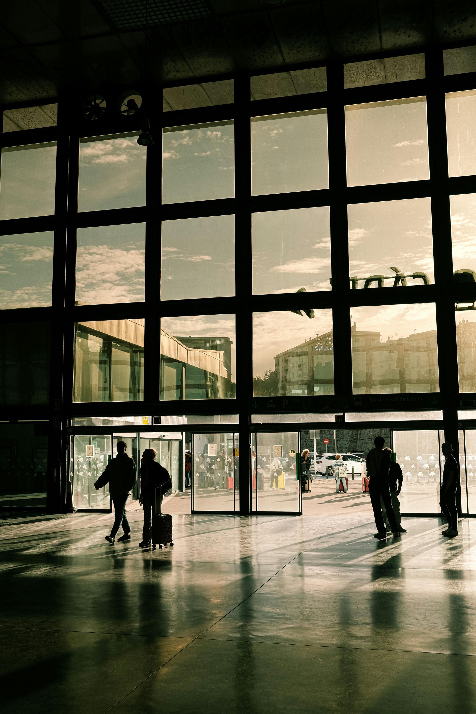 People walking through glass doors of a building. Sunlight shines through the windows; reflections on the floor.