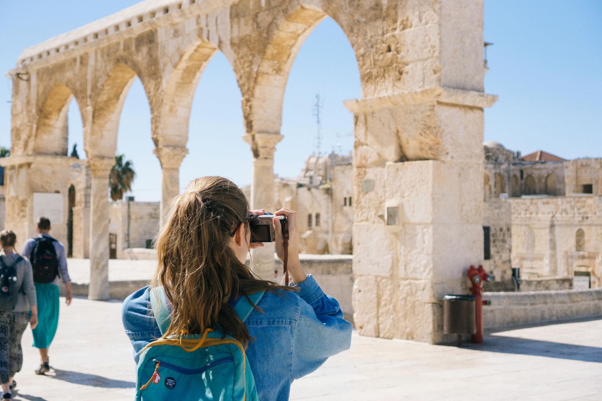 Woman taking a photo under arched stone structures; people walking in the background, sunny day.
