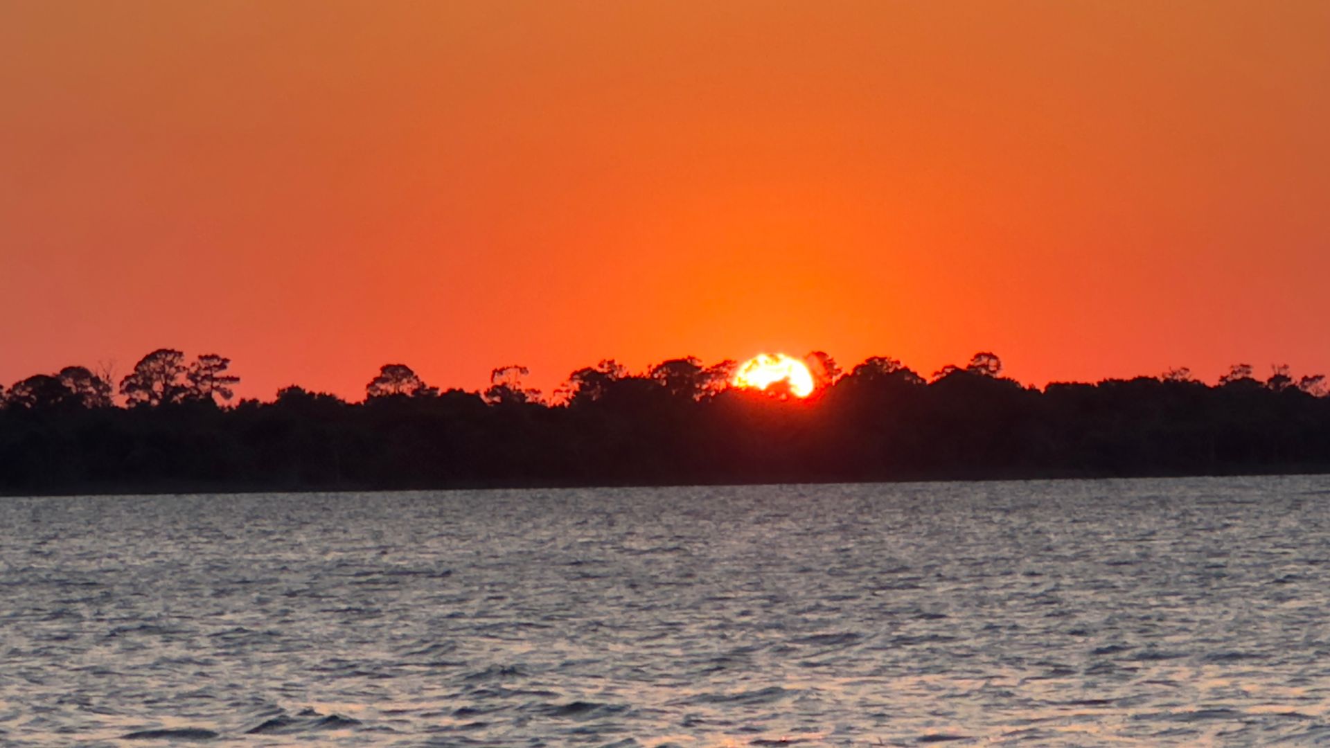 Sunset over water, with trees silhouetted against the bright orange and red sky.