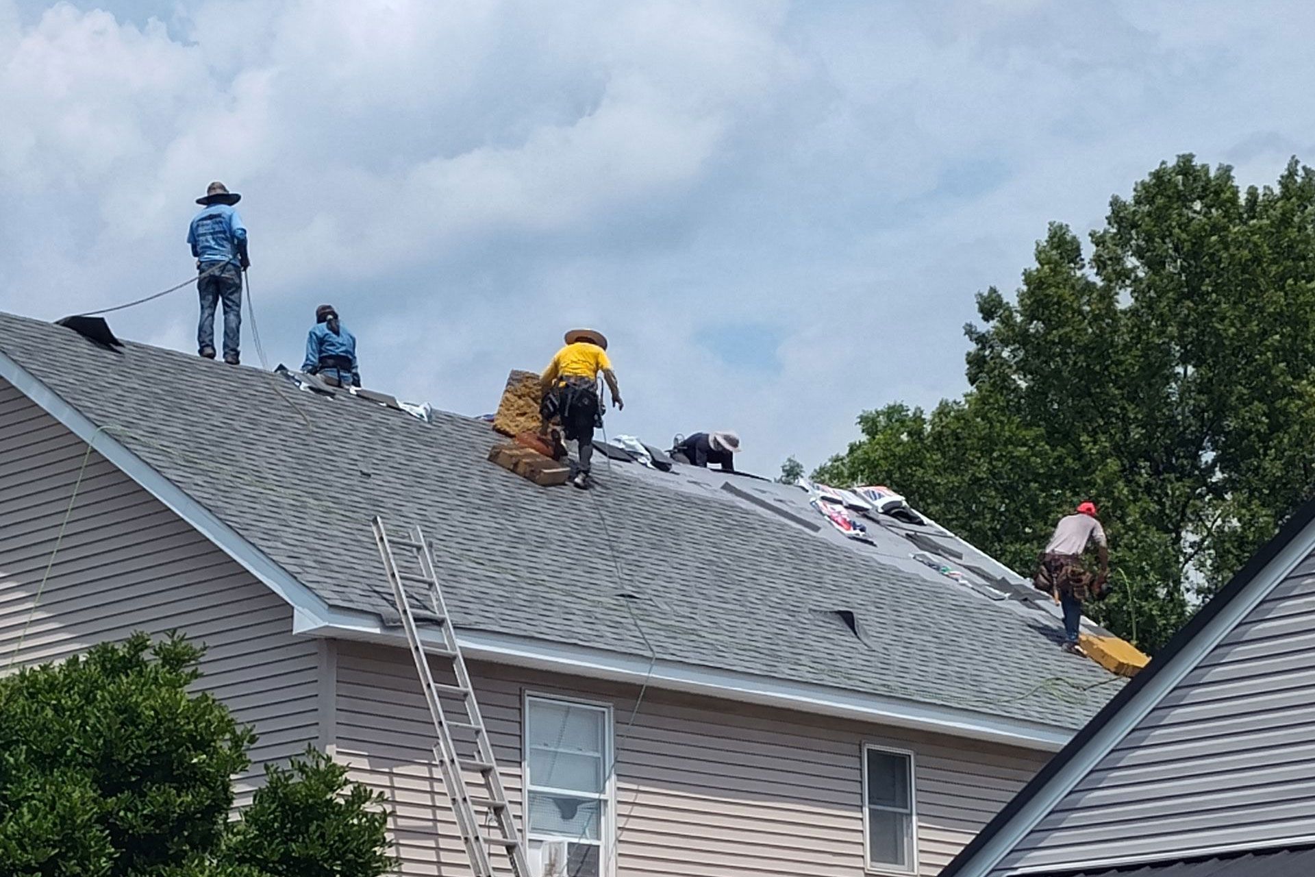 Roofers working on a residential roof, wearing safety gear and using tools. Cloudy sky and trees in background.