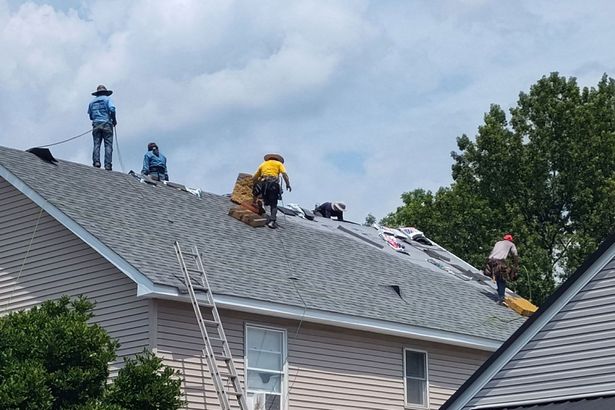 Roofers working on a residential roof, wearing safety gear and using tools. Cloudy sky and trees in background.
