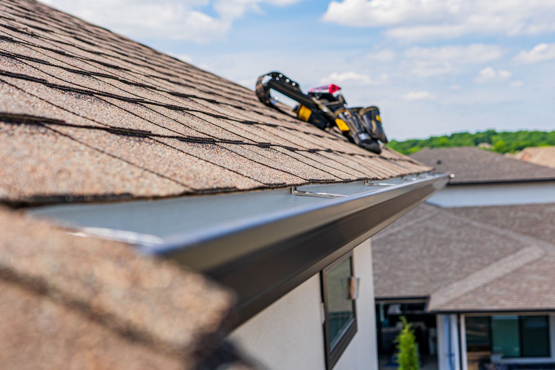 Gutter installation on a brown shingle roof. Tools sit on roof edge, overlooking a sunny suburban landscape.