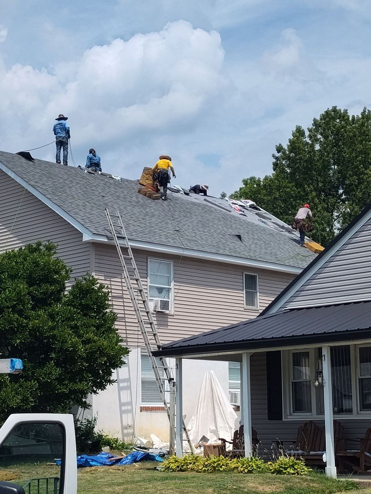 Roofers replacing shingles on a two-story house, working on a sunny day. A ladder leans against the building.