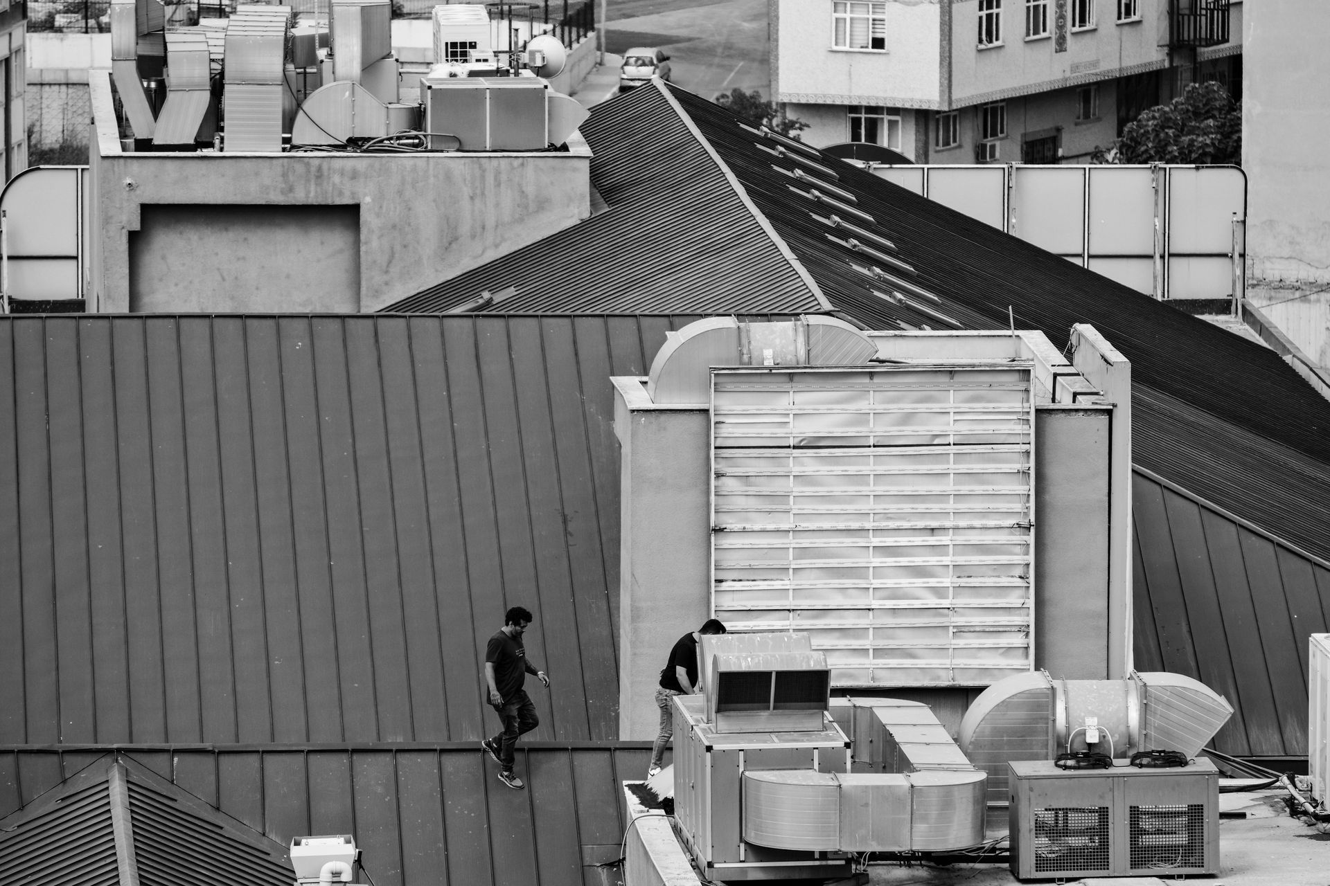 Two people on a metal rooftop with ventilation systems. City buildings in the background. Black and white.
