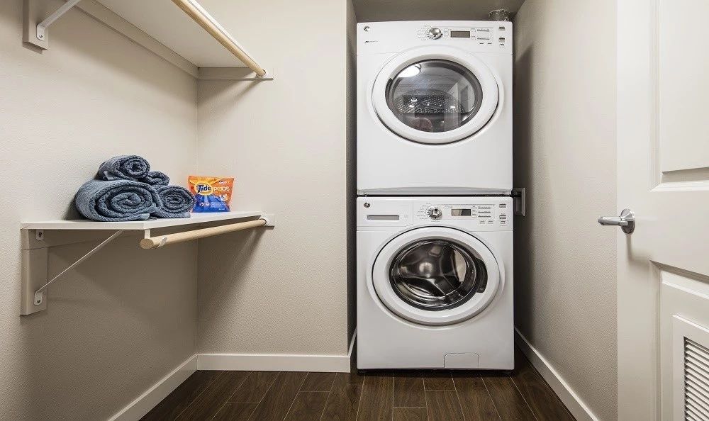 Laundry room with a stacked washer and dryer, shelf with towels and laundry detergent.