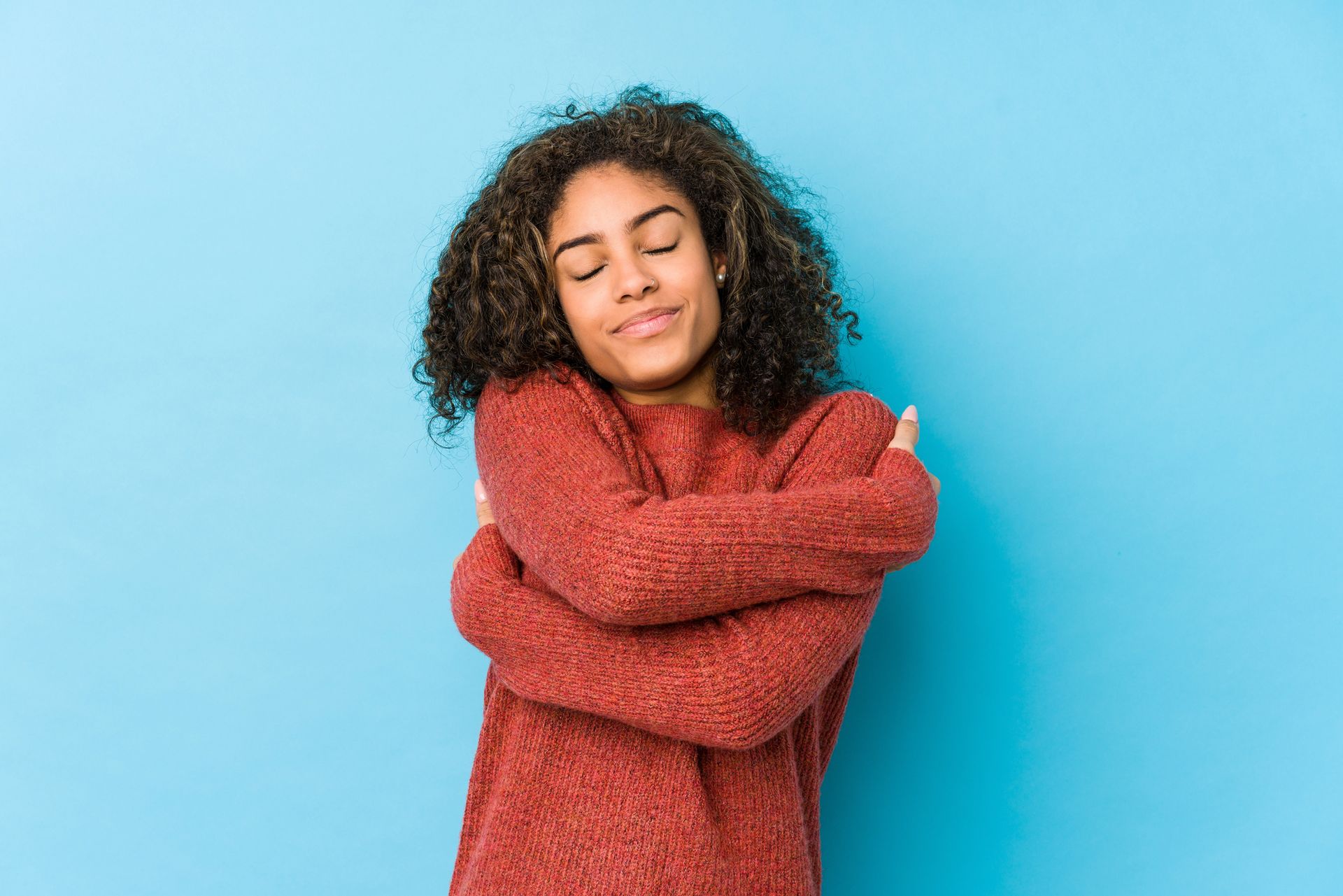 Woman with curly hair in a red sweater hugging herself, eyes closed, against a blue background.