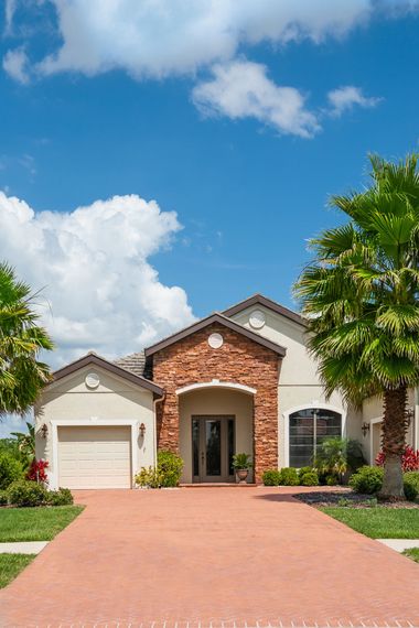 Beige house with a red brick driveway and a blue sky with fluffy clouds.