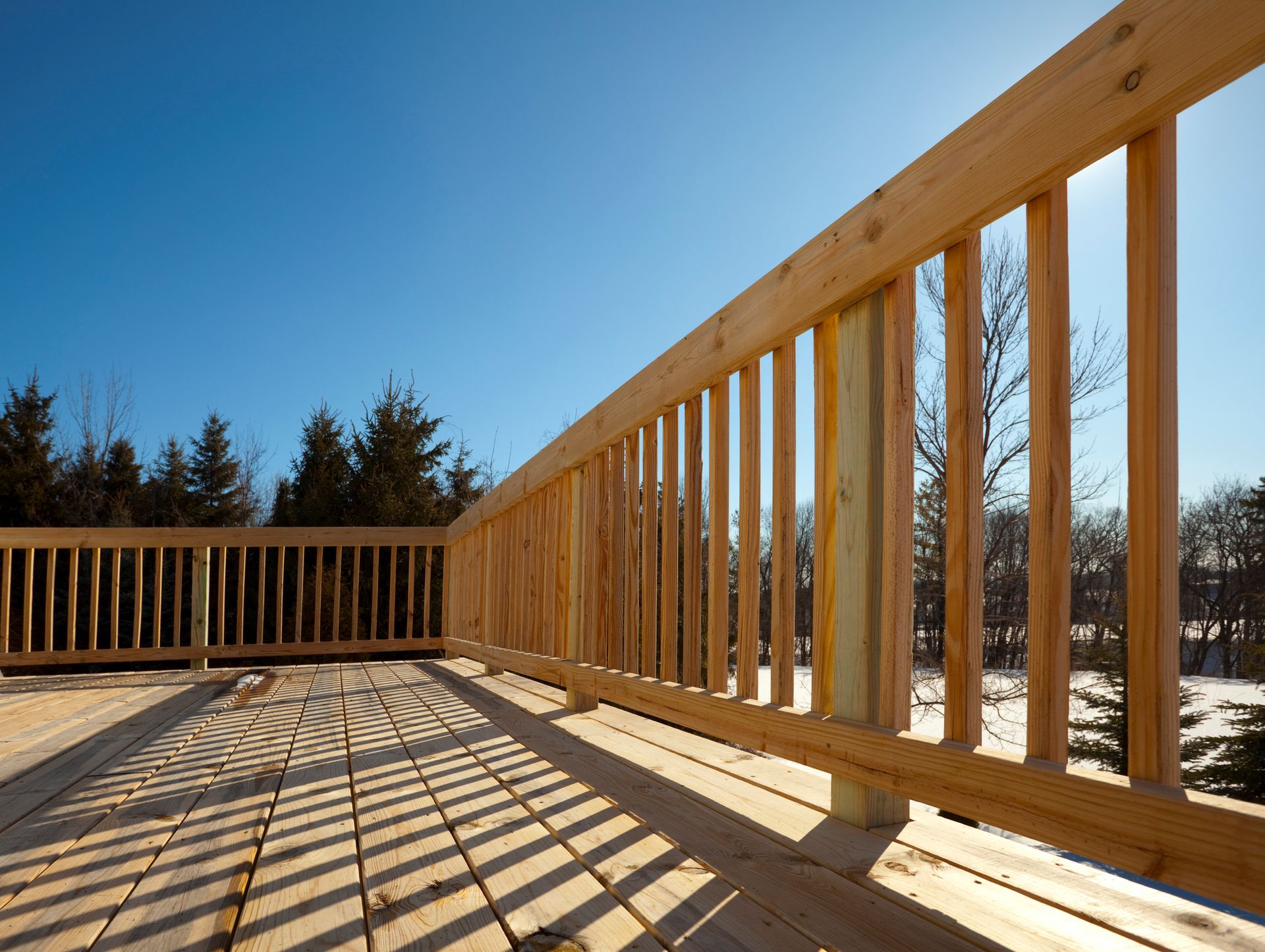 Wooden deck with railing, shadows on the planks, bright blue sky, trees in the background.