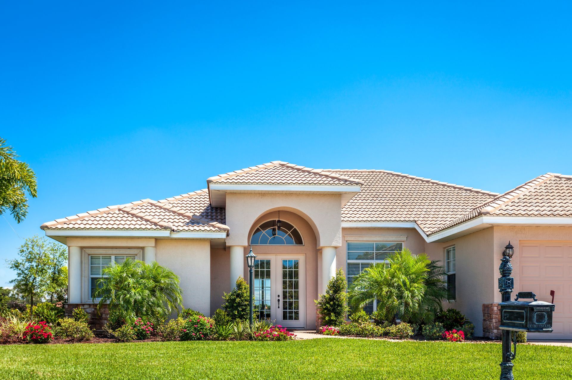 Beige stucco house with light tile roof under blue sky, front lawn with green grass and landscaping.