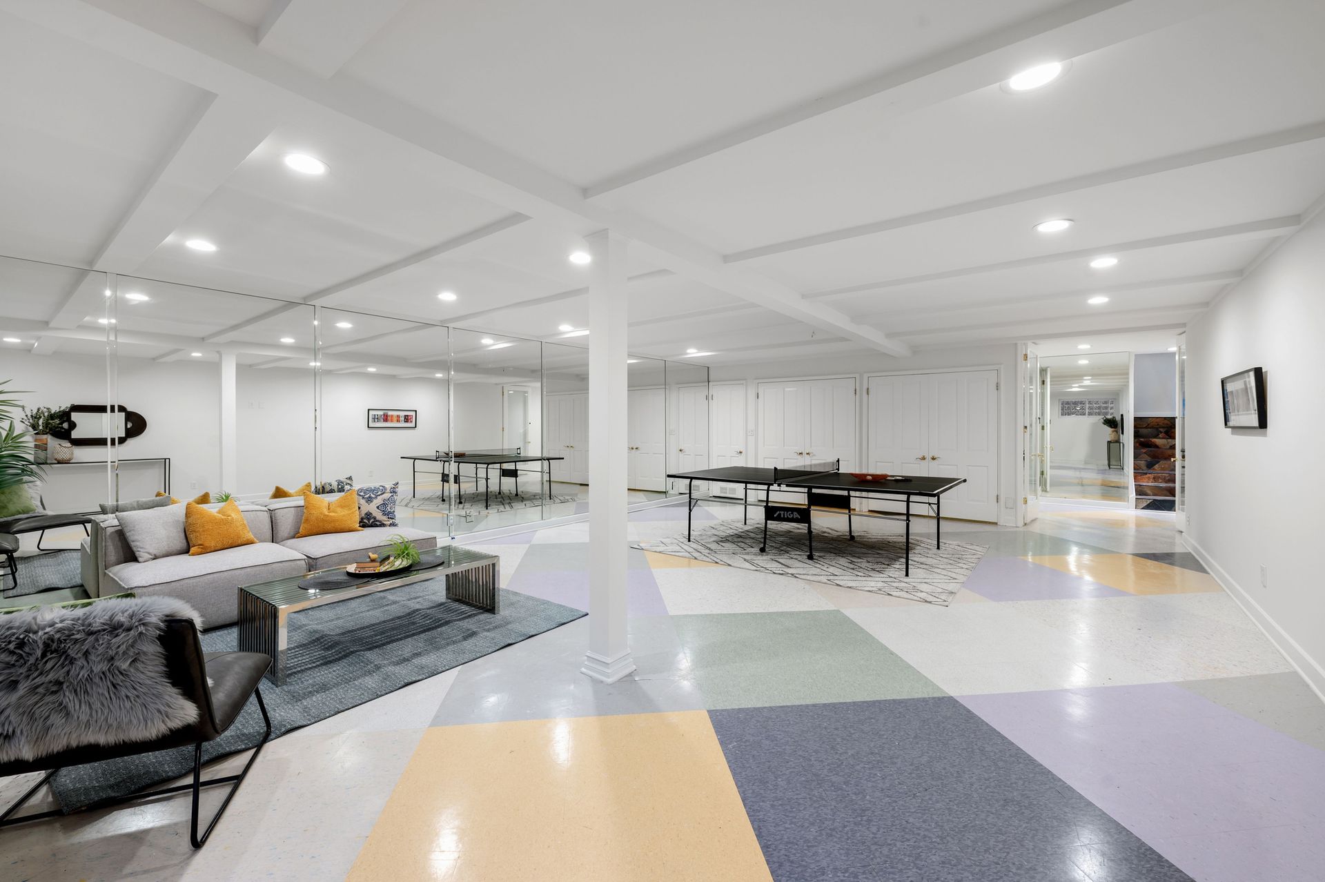 Spacious, bright basement with colorful geometric flooring, ping pong tables, seating area, and white walls. Spacious, bright basement with colorful geometric flooring, ping pong tables, seating area, and white walls.