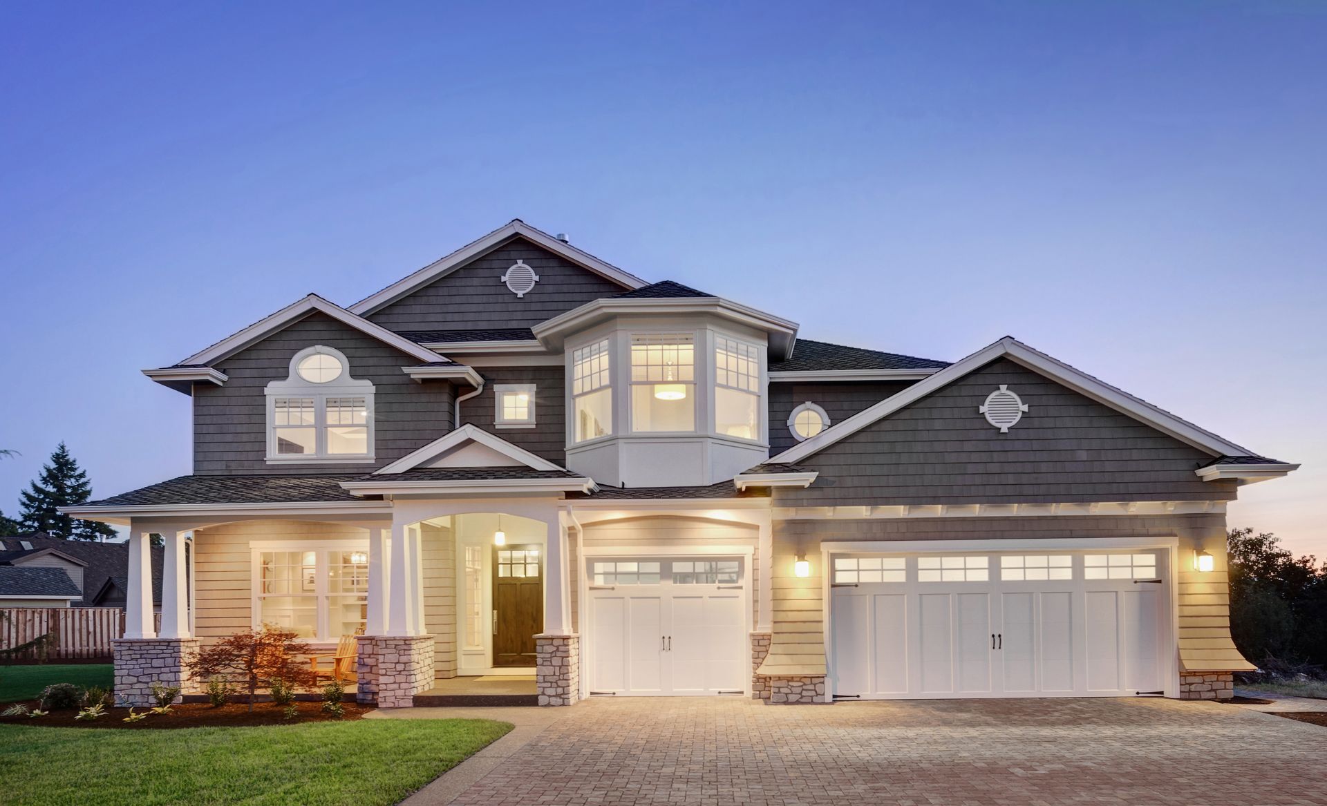 Two-story house at dusk, featuring a bay window, two-car garage, and a long driveway; exterior lights are on.