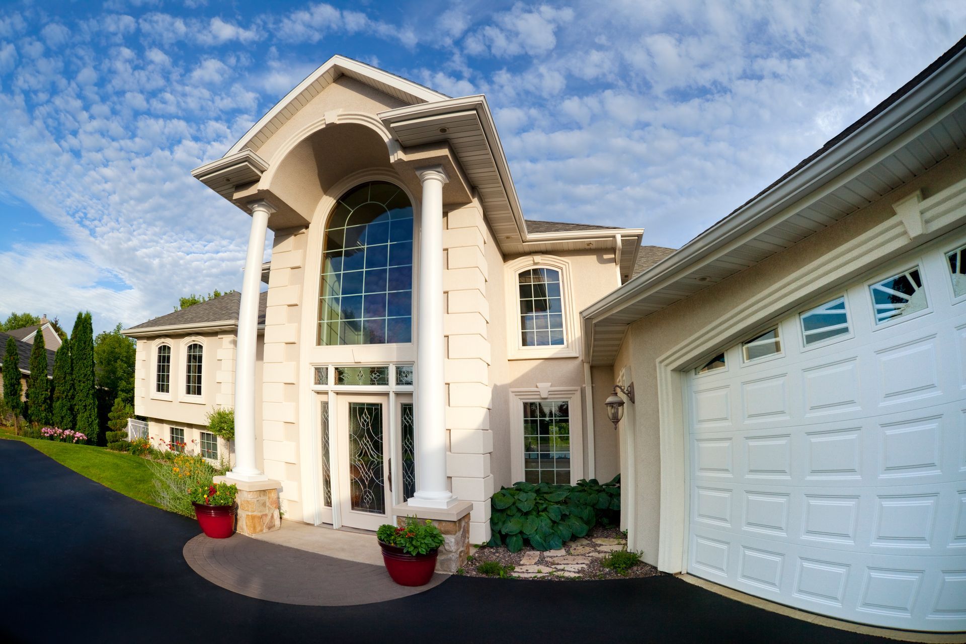 Beige two-story house with tall white columns, arched front window, and attached garage. Black driveway and blue sky.