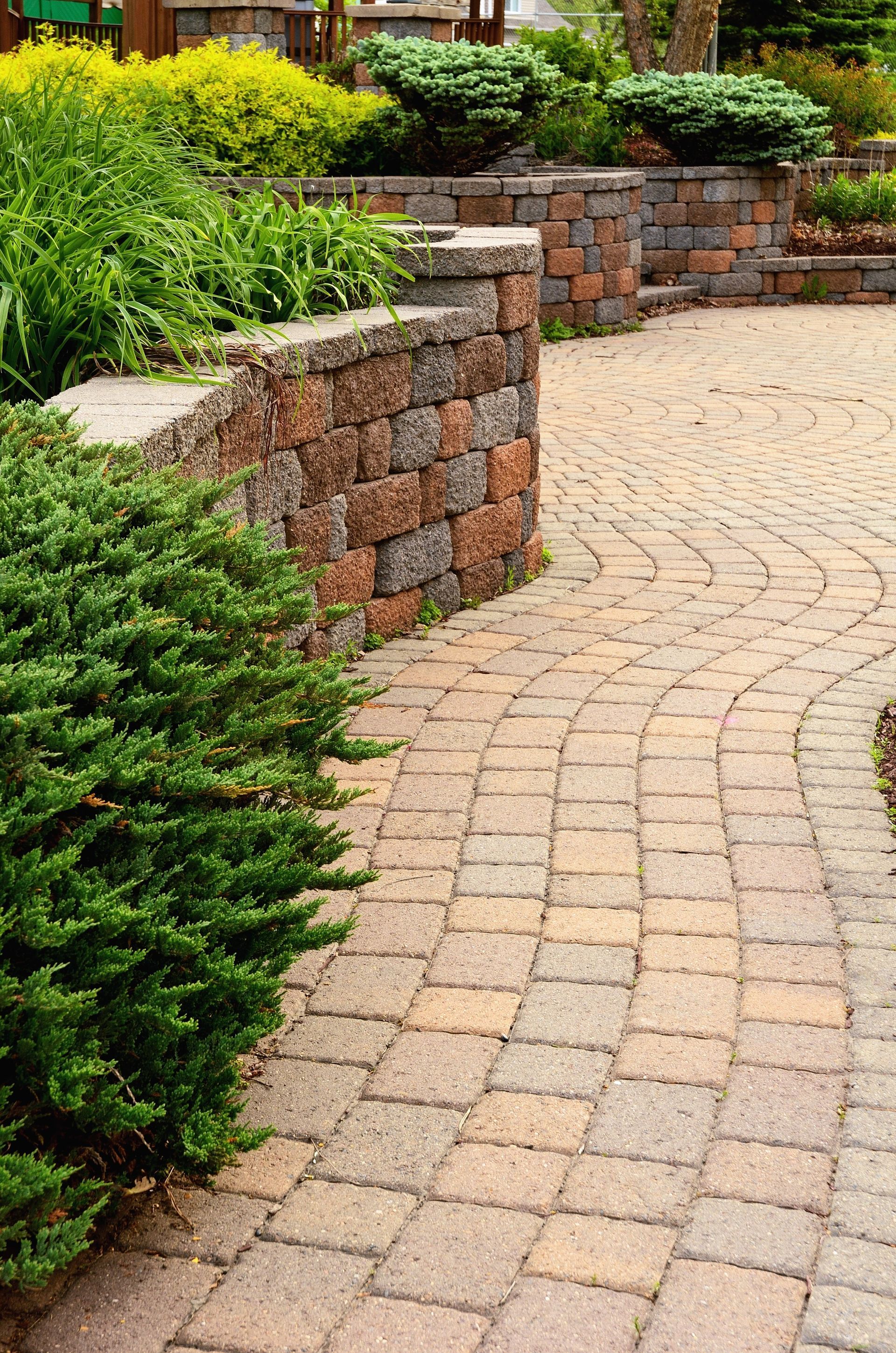 Brick pathway curving past a stone retaining wall with lush greenery.