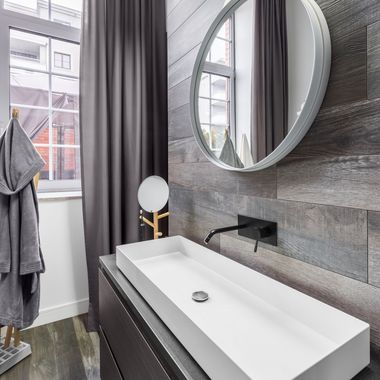 Bathroom with a rectangular white sink, black faucet, round mirror, and gray wall paneling. A gray robe hangs on a wooden stand.