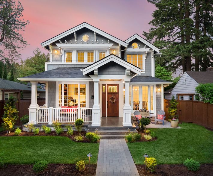 Two-story gray house with white trim, porch, and a brick walkway. Green lawn and a pink sunset sky.