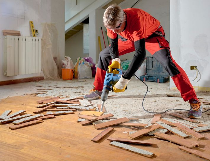 Person in red work clothes using a jackhammer to remove floorboards in a room.