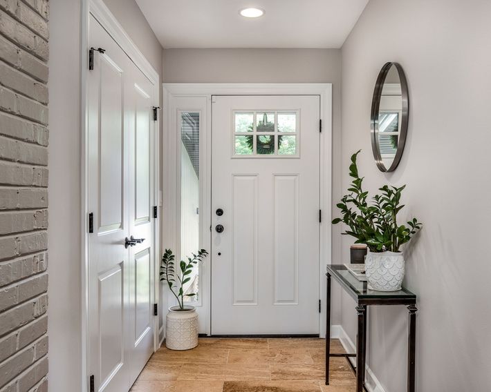 White entryway with a front door, side window, and decorative table with plants and a mirror.