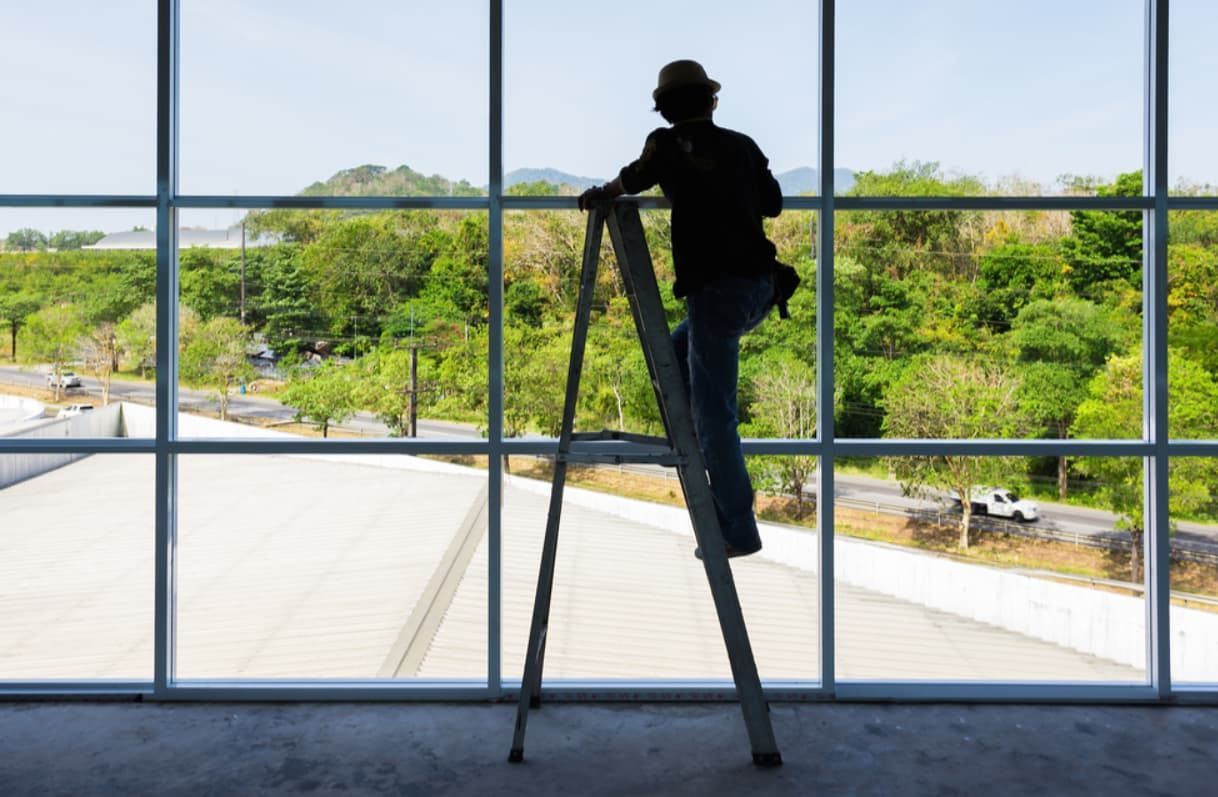 A Man Is Standing On A Ladder Looking Out A Window — Northern Glazing Glass & Aluminium In Smithfield, QLD