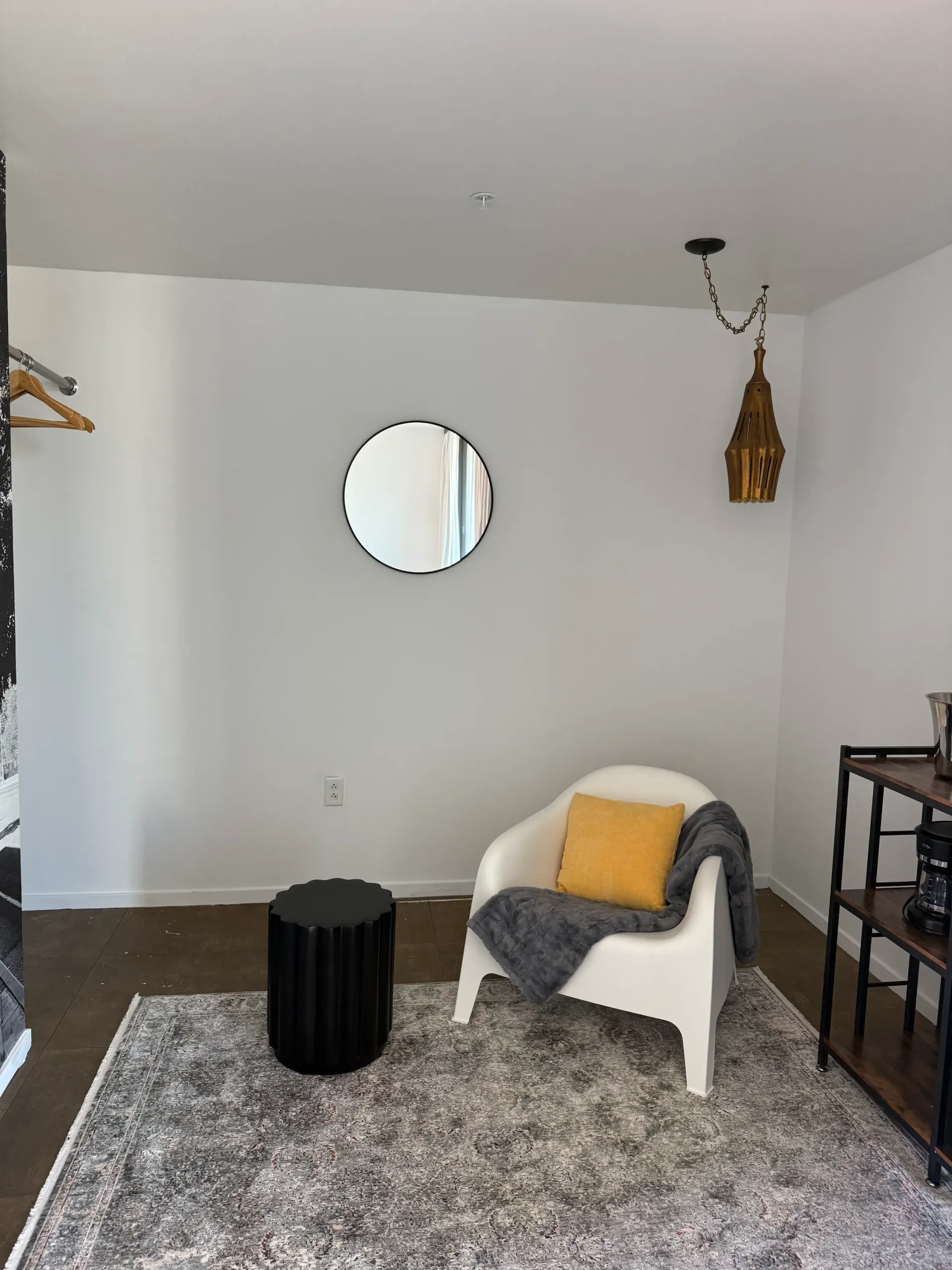 A cozy reading nook with a white armchair, yellow pillow, gray throw blanket, black side table, rug, mirror, and lamp.