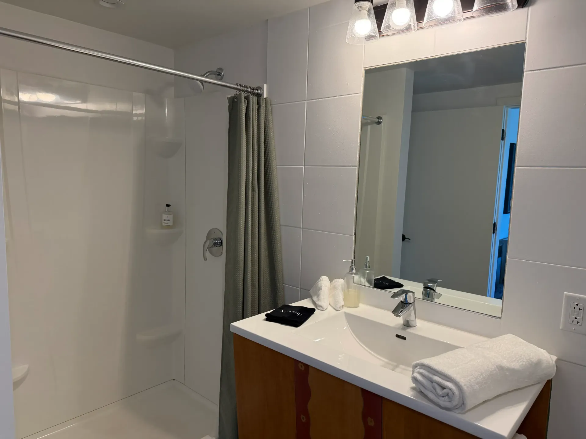 A hotel bathroom with a white sink, wood vanity, mirror with a light fixture above, and a shower with a gray curtain.