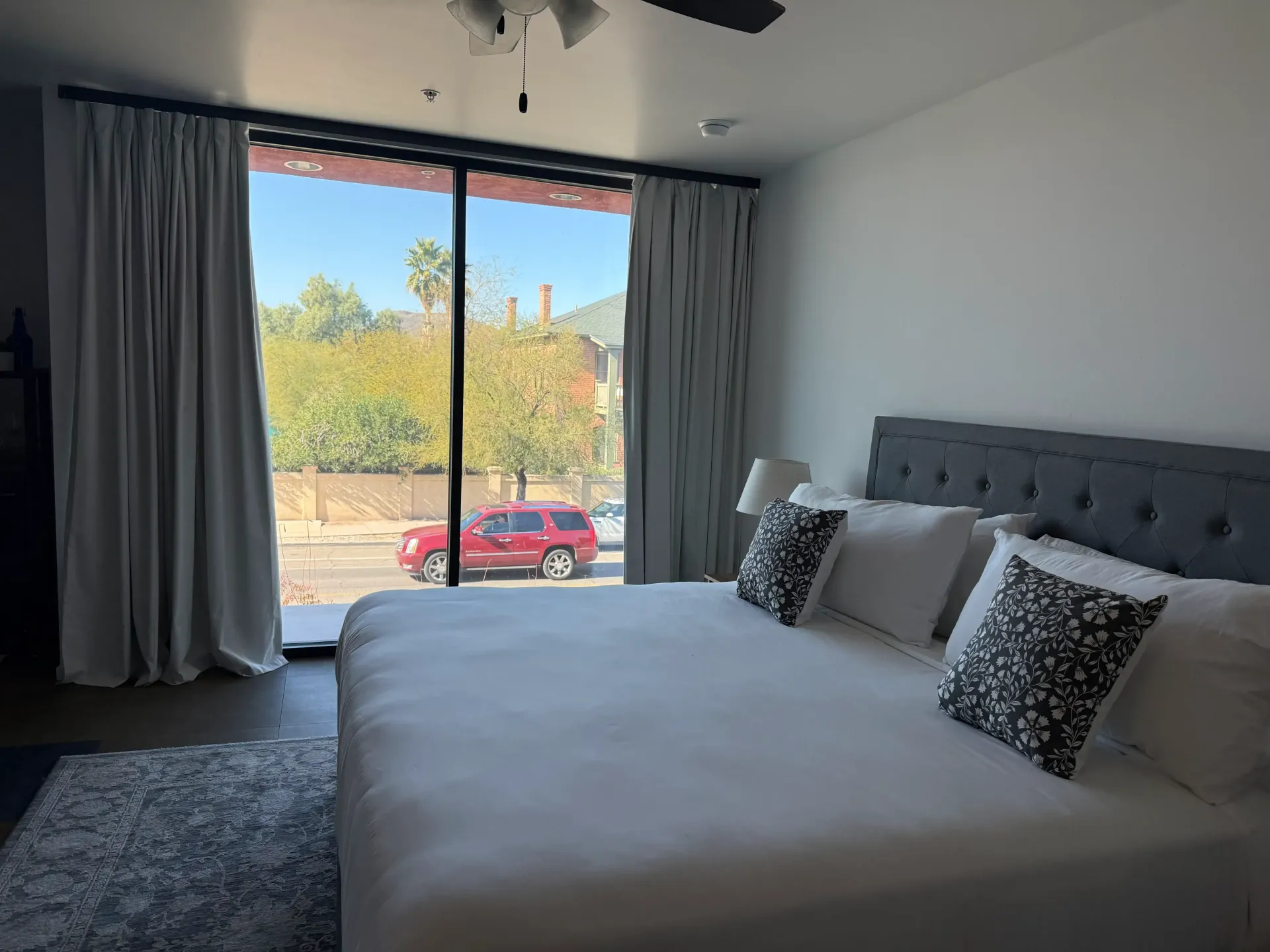 A bright hotel room featuring a large bed with white linens and patterned throw pillows, next to floor-to-ceiling windows.