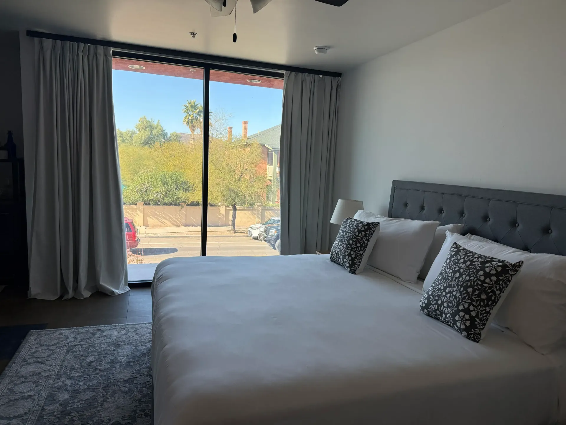 A bright hotel room featuring a king bed with white linens, patterned pillows, and a large sliding glass door with curtains.