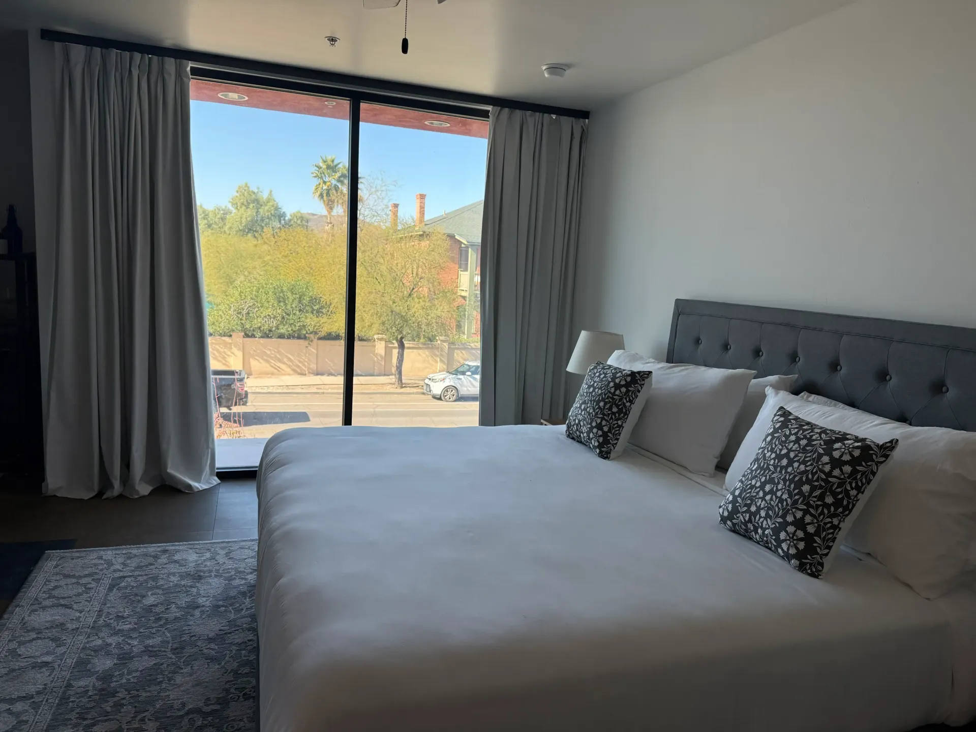 A bright bedroom with a king-sized bed, white linens, patterned pillows, and a large window overlooking a sunny street.