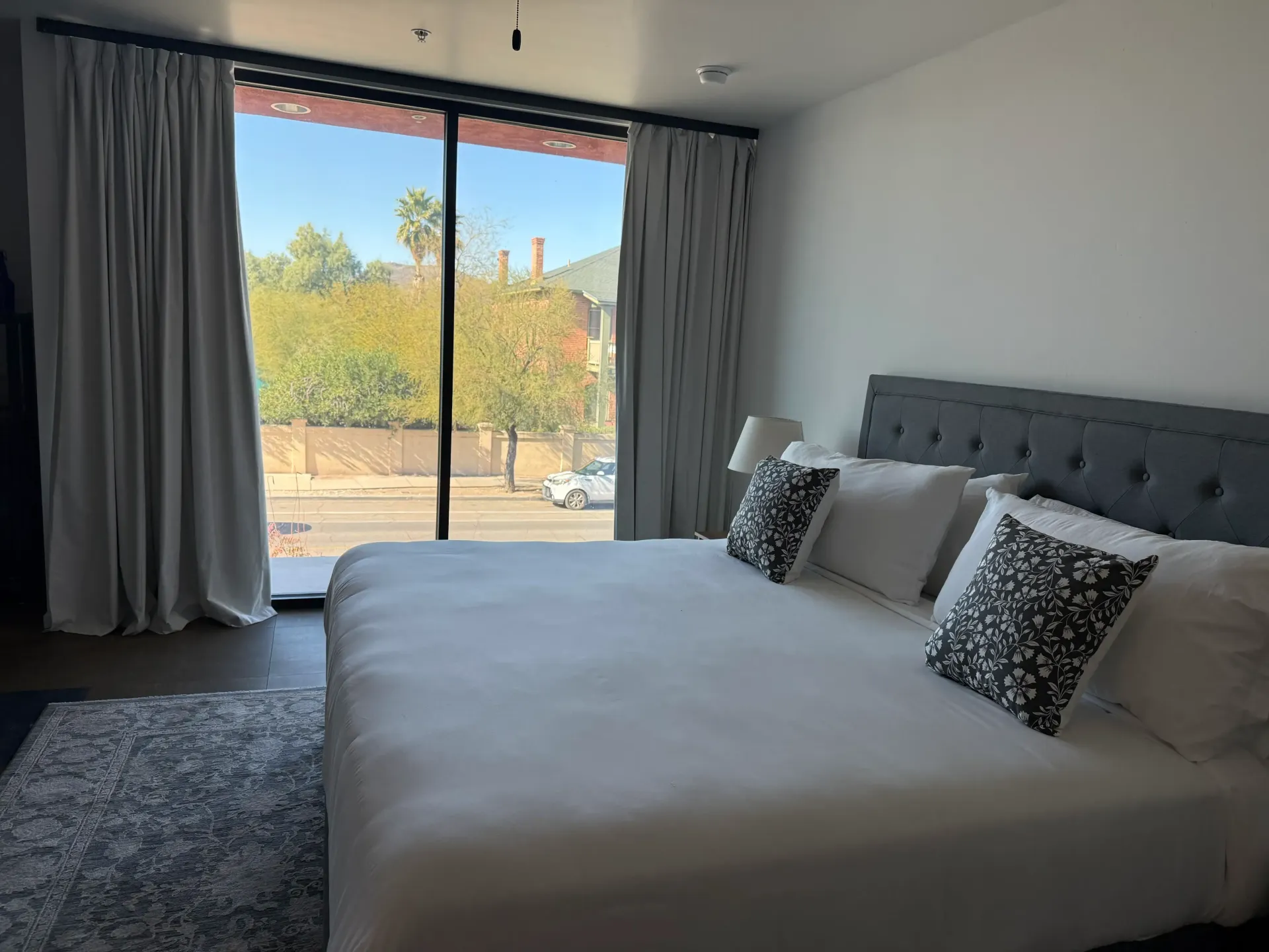 A bedroom with a king-sized bed, white linens, patterned pillows, a grey tufted headboard, and floor-to-ceiling curtains.