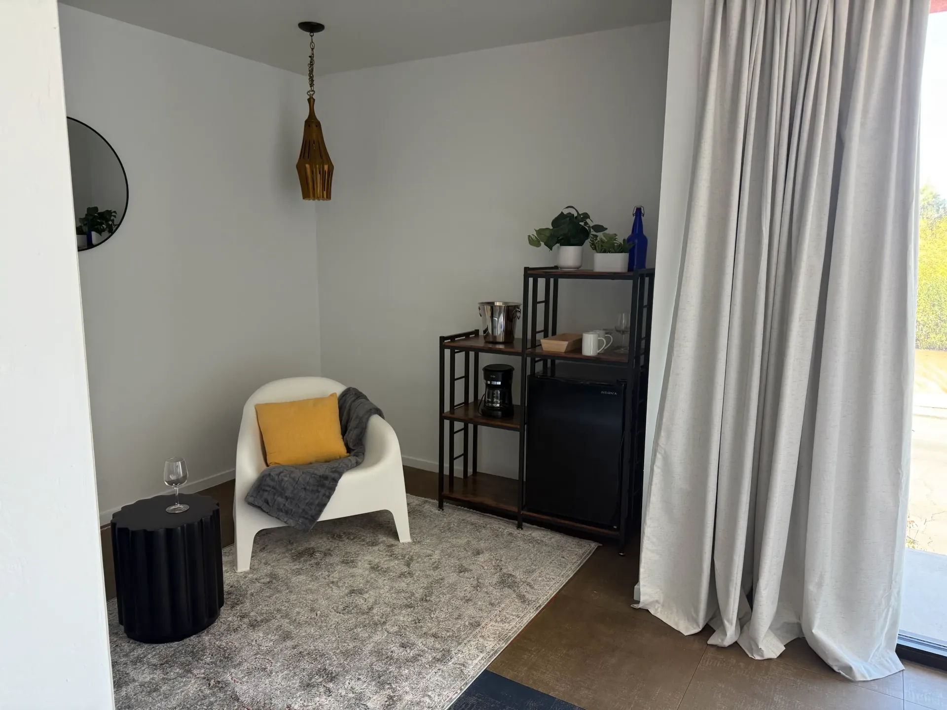 A cozy corner with a white armchair, yellow pillow, fuzzy gray rug, black side table, and a shelving unit with a plant.