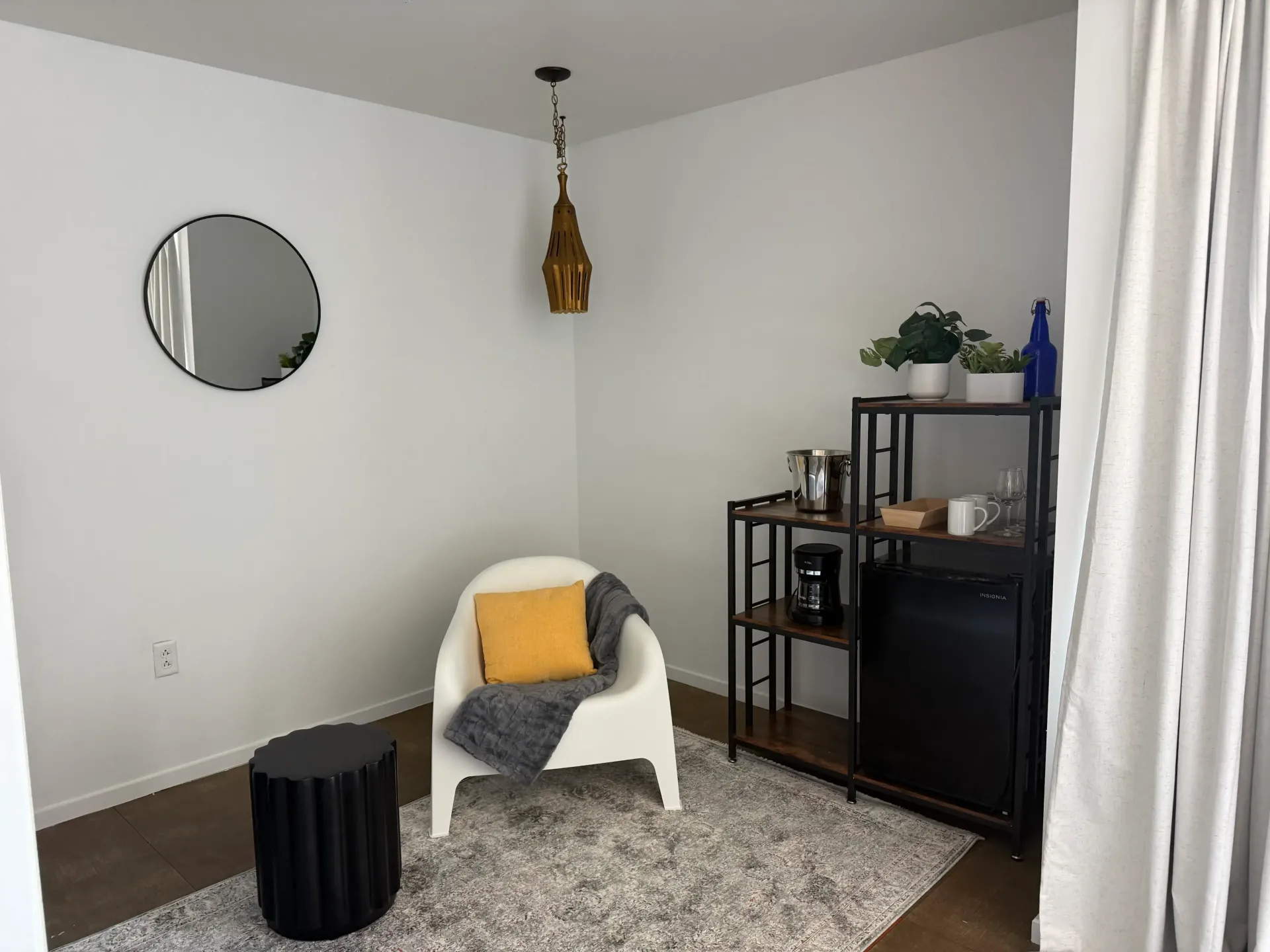 A cozy corner featuring a white armchair with a yellow pillow, a side table, a shelving unit, and a round wall mirror.