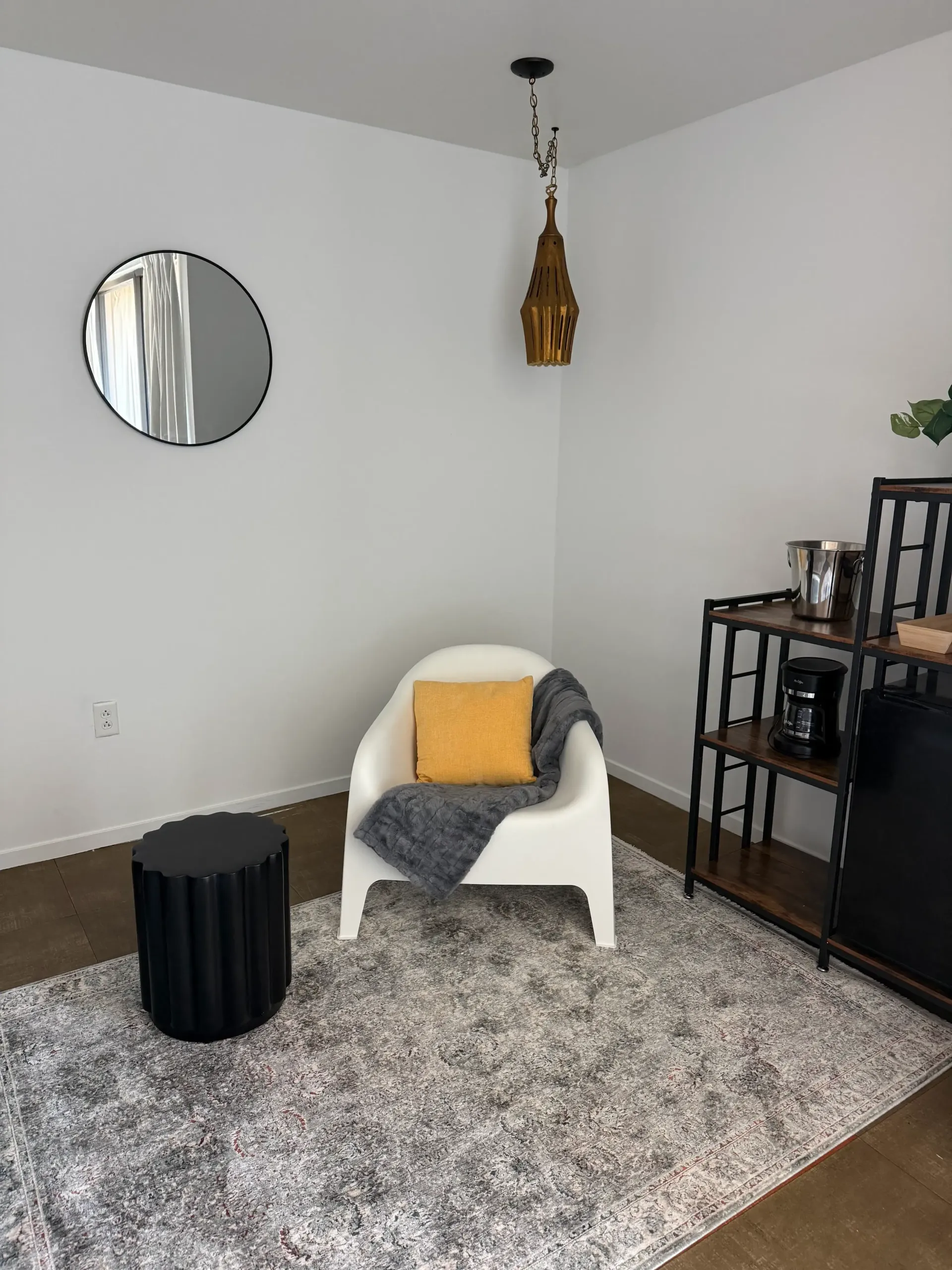 A cozy seating nook with a white armchair, a yellow pillow, a black side table, and a decorative pendant light on a rug.