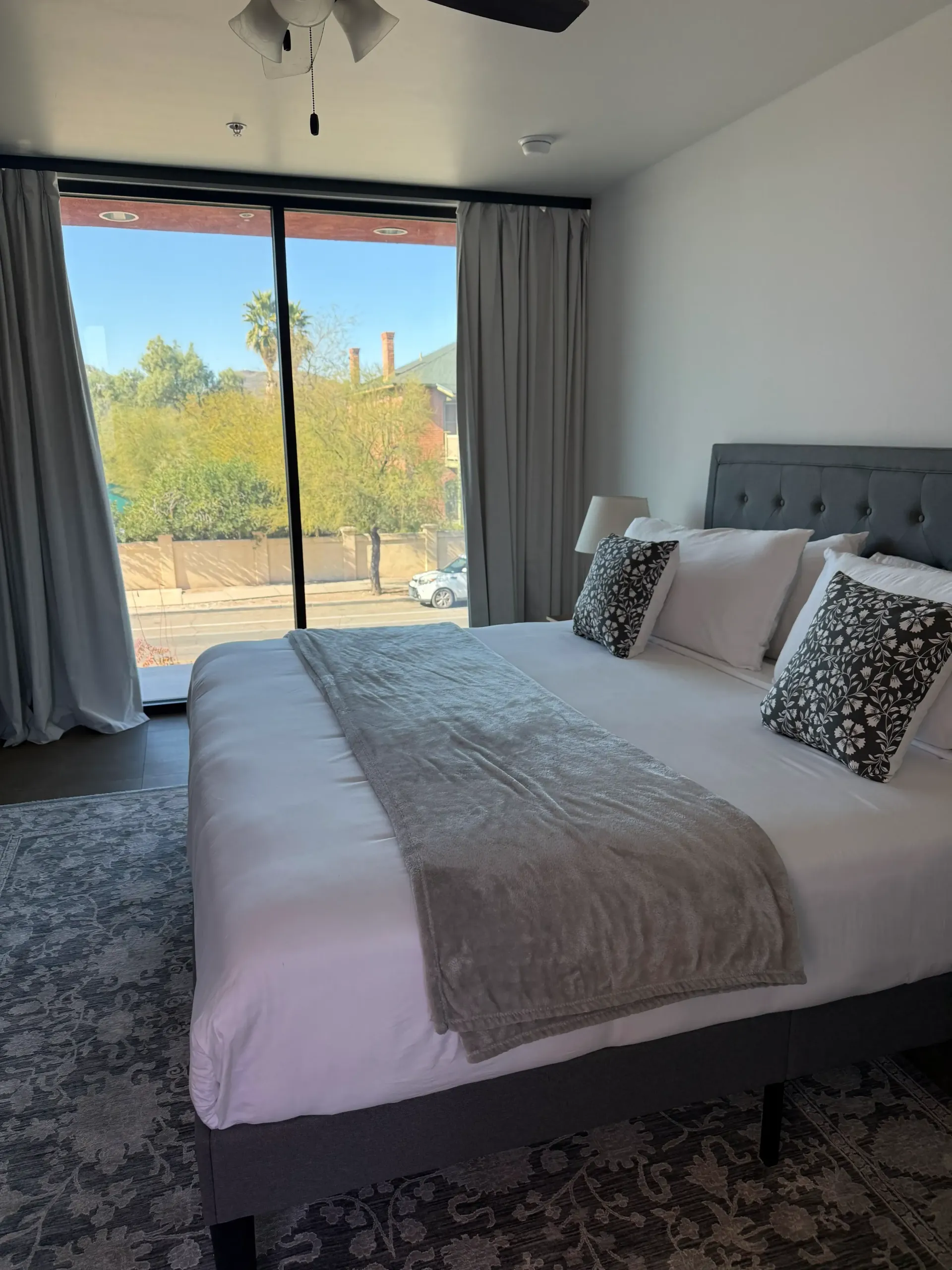 A bedroom with a king-sized bed, grey tufted headboard, and large floor-to-ceiling windows looking out onto a sunny view.