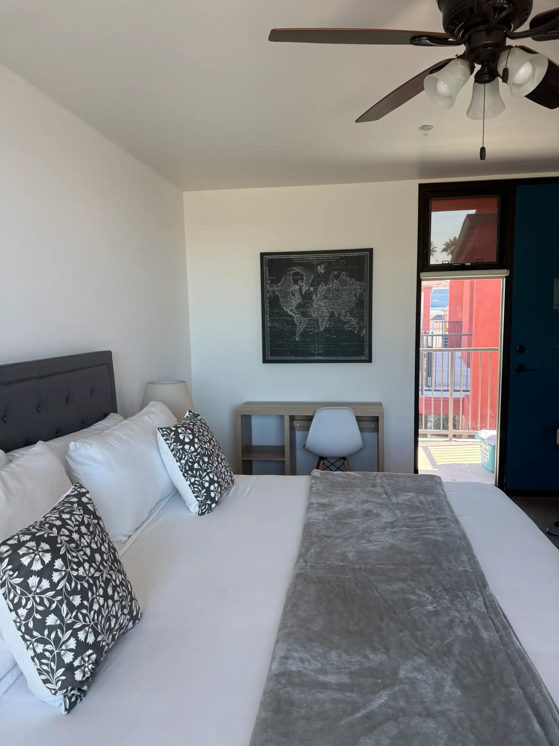 A bedroom featuring a bed with white bedding, a grey patterned throw, a dark tufted headboard, a desk, and a wall map.