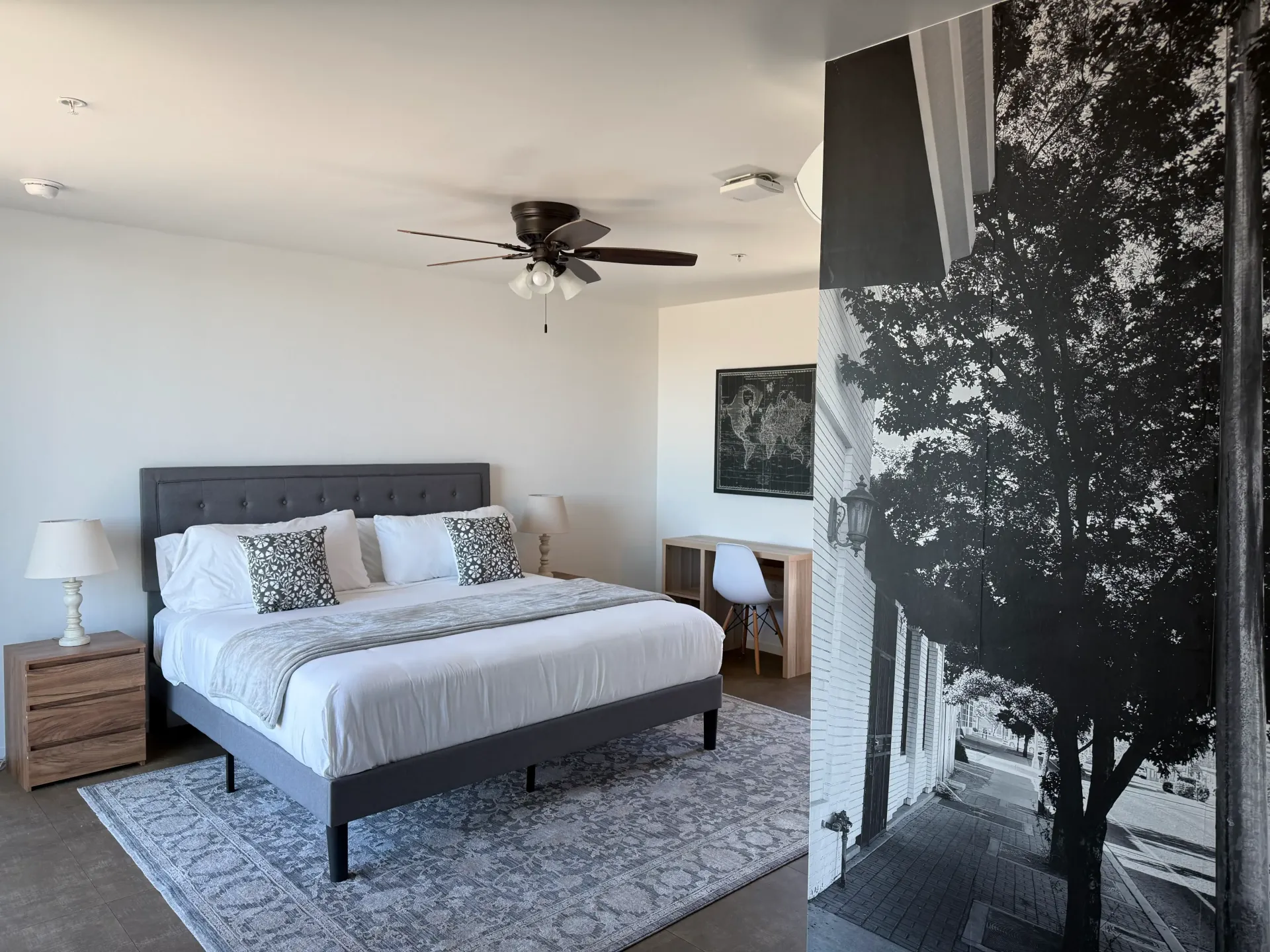 A modern bedroom with a grey tufted bed, white bedding, a patterned rug, and a large black-and-white tree mural on the wall.