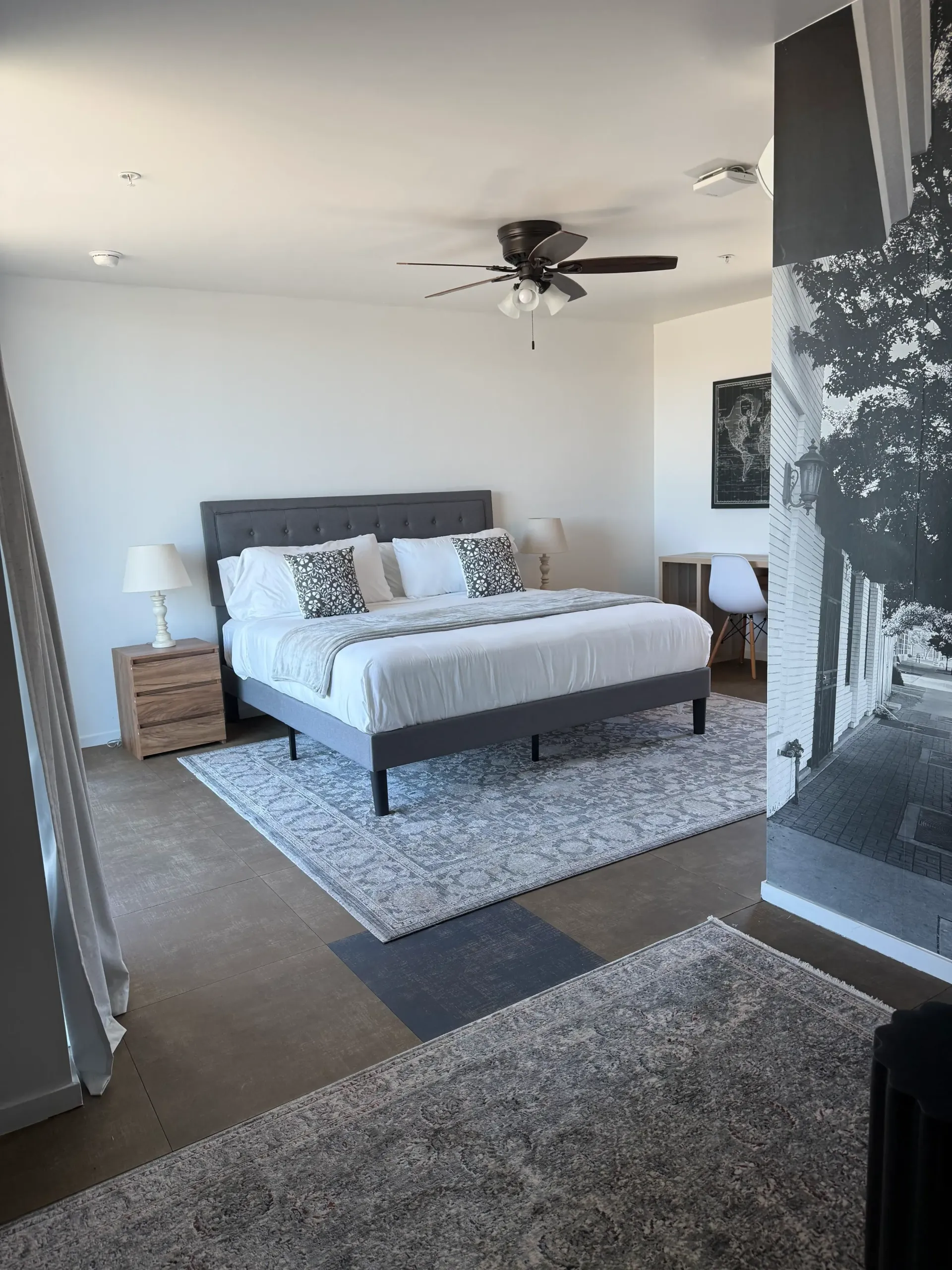 A modern bedroom with a grey tufted bed, white bedding, two matching nightstands, and a large black-and-white mural wall.