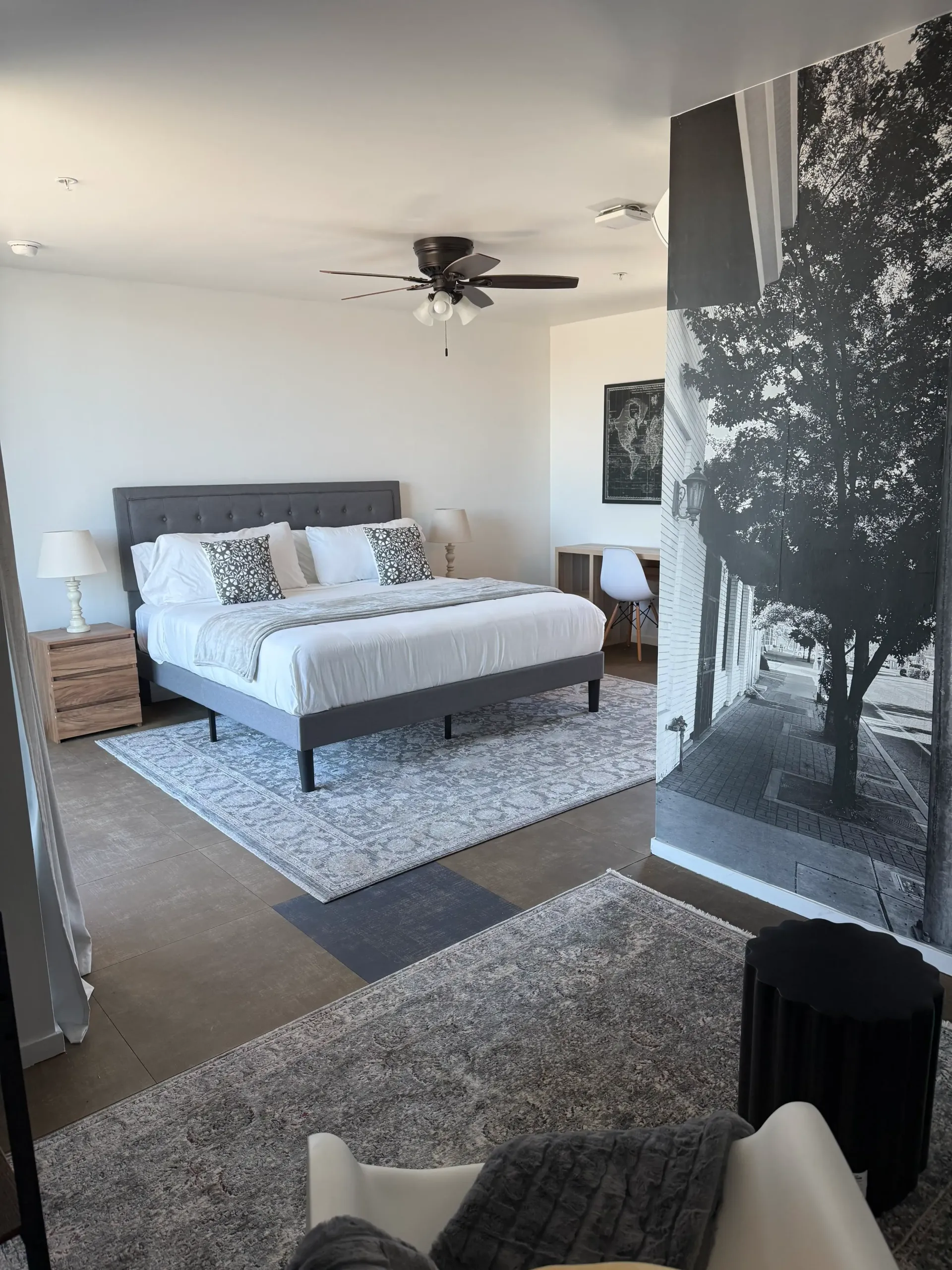 A spacious, modern bedroom with a grey tufted bed, shag rugs, and a large black-and-white tree mural on the accent wall.